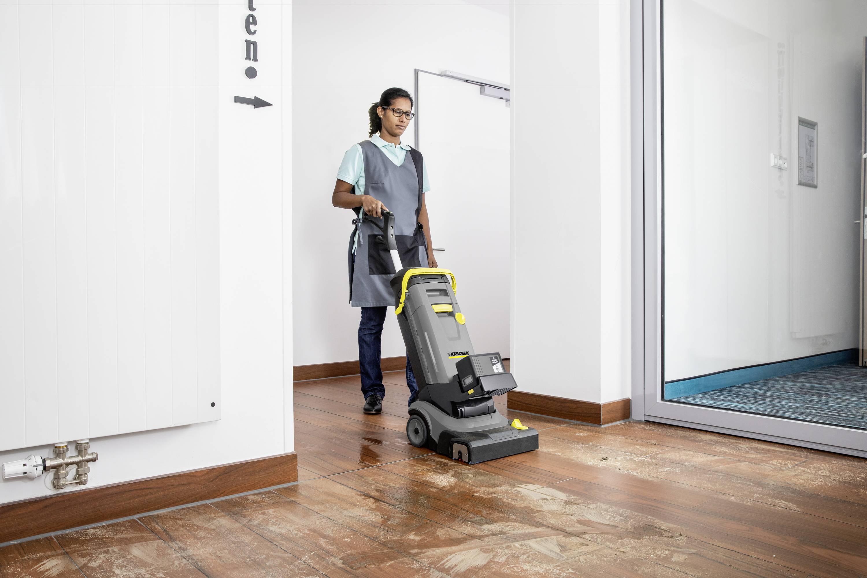 A person is cleaning a wooden floor with a grey floor cleaning machine in a bright room with a glass wall on the right.