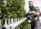 A person with a beard is trimming a dense, green hedge using an electric hedge trimmer. They are wearing safety glasses and gloves.