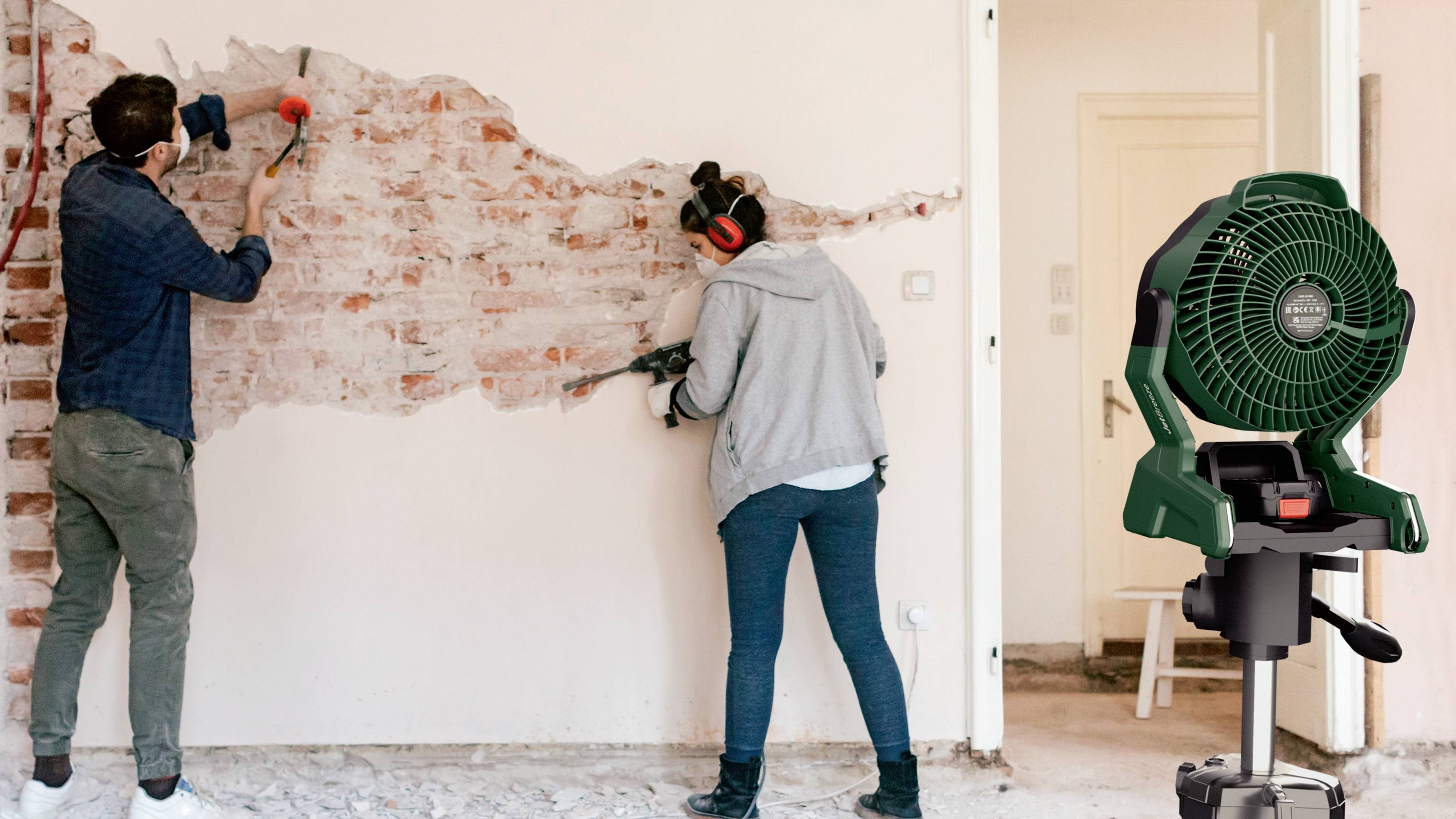 Two people are removing plaster from a brick wall using tools. A green fan is situated to the right, presumably for ventilation.