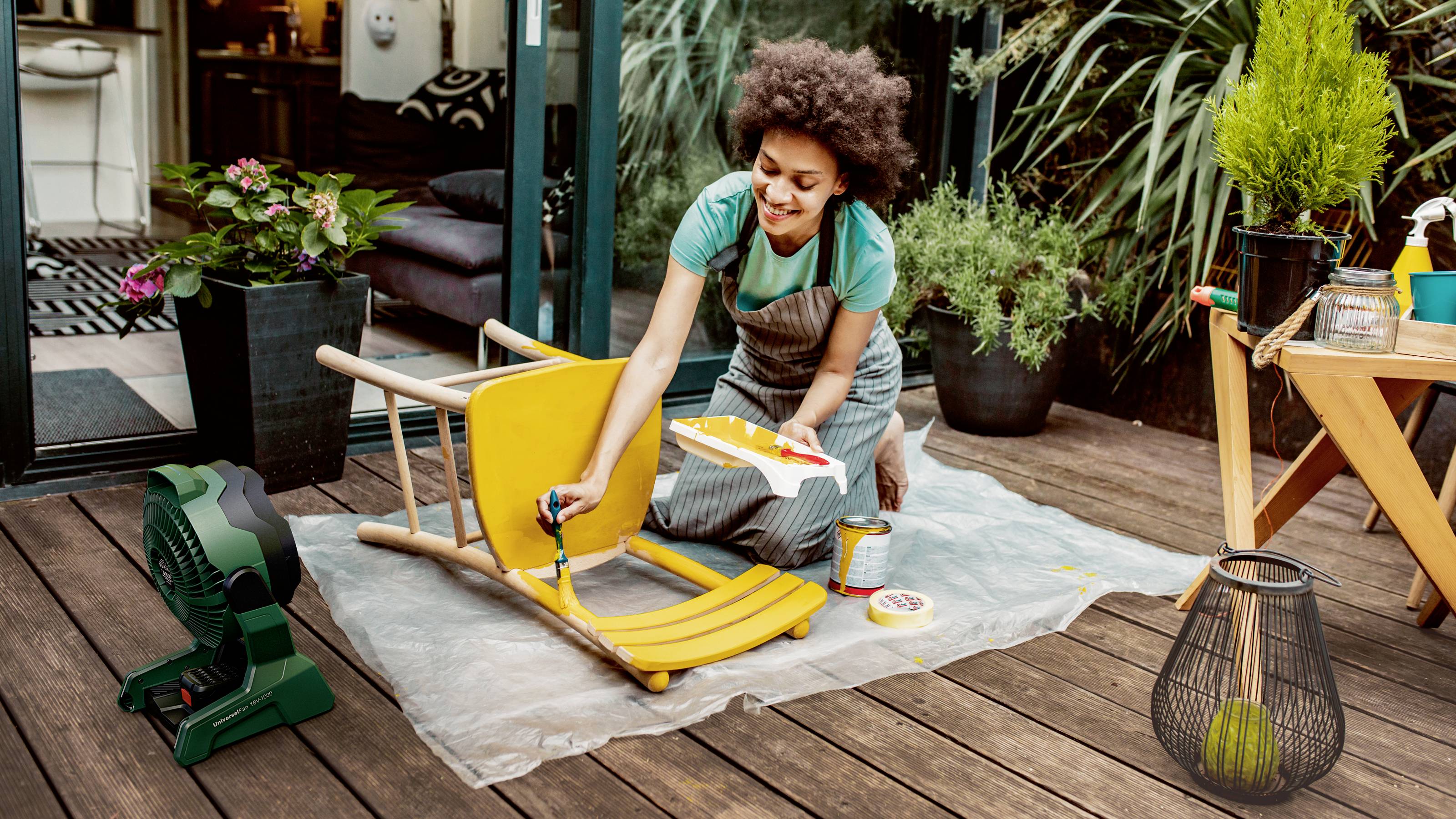 A person is painting a yellow chair on a terrace. Plants and tools are visible in the background.