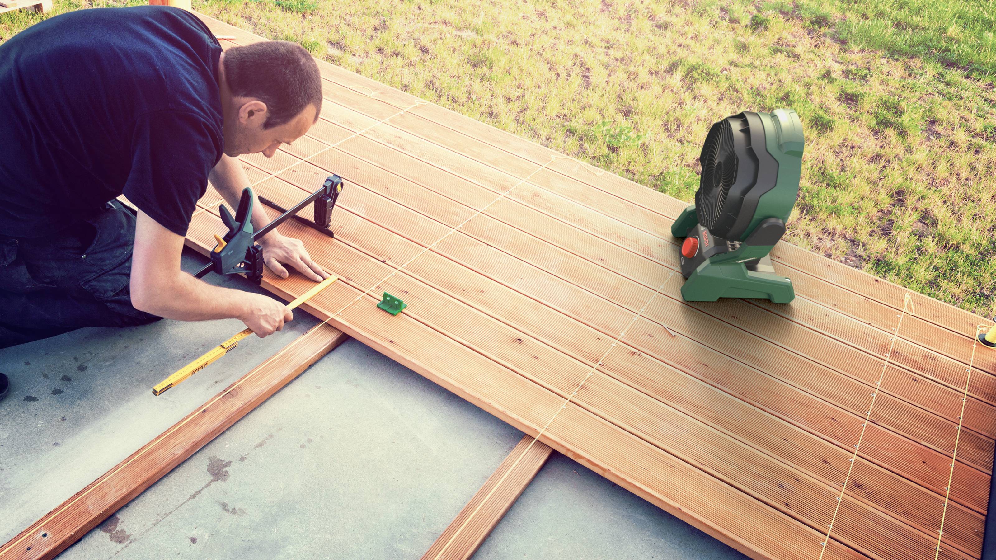 A man is working in the garden woodworking, measuring wooden battens on a terrace with tools in daylight.