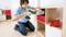 A man is assembling a shelving unit, kneeling on the floor and using an electric drill. The shelving unit has red and white drawers.