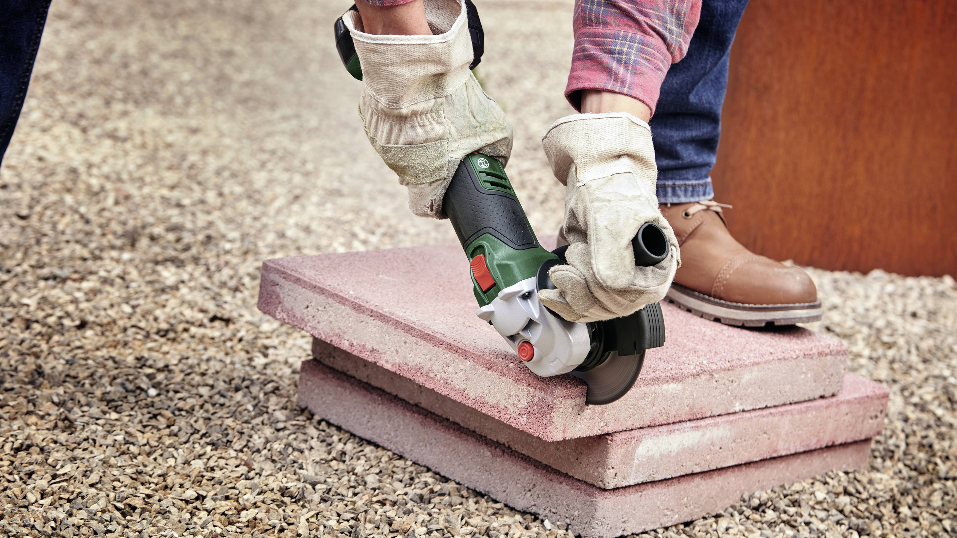 A person wearing gloves is cutting a rectangular stone slab with an angle grinder while standing on a gravel surface.