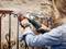 A person is grinding a metal fence with an angle grinder. They are wearing work gloves and a denim jacket. A wall is visible in the background.
