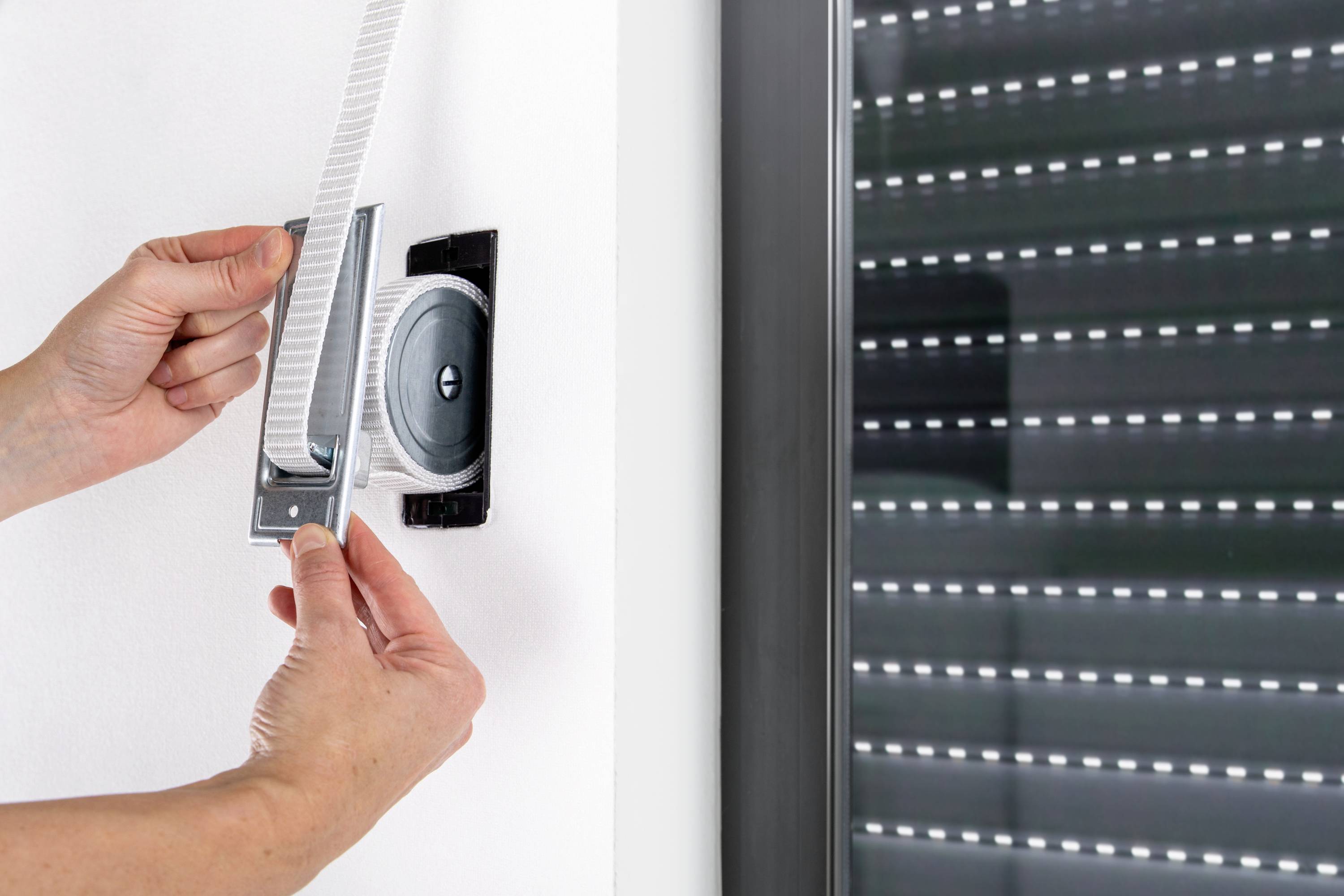 A person pulls on a roller blind cord next to a closed slatted window.