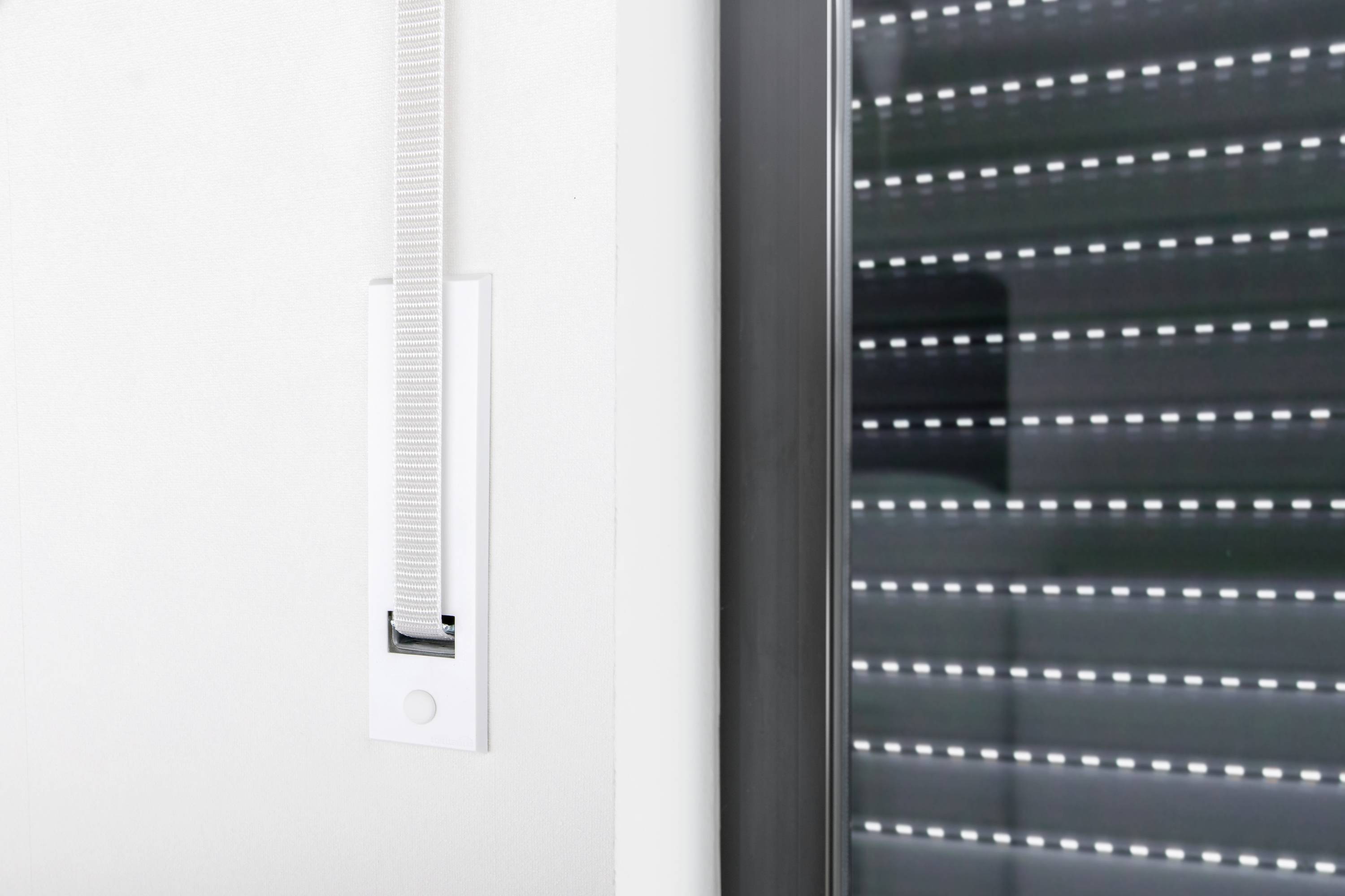 An automated roller blind opens at a large window in a modern room. The motor and strap are visible.