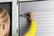 A person in a yellow jumper is repairing a grey roller shutter by tightening a screw with a screwdriver.