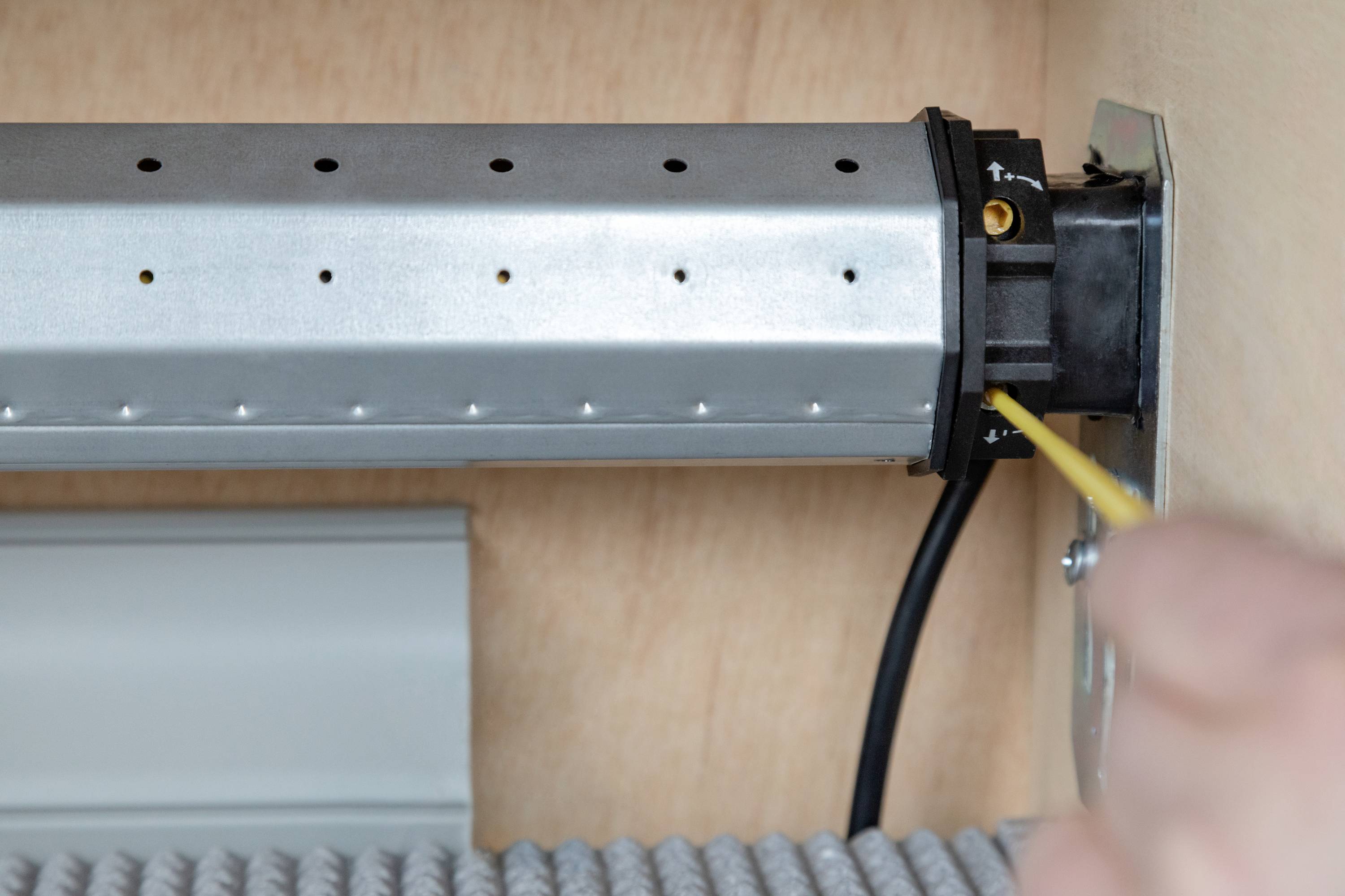Close-up of a roller shutter motor in a box. A hand is pointing to the motor's control unit with a tool. Cables are visible.