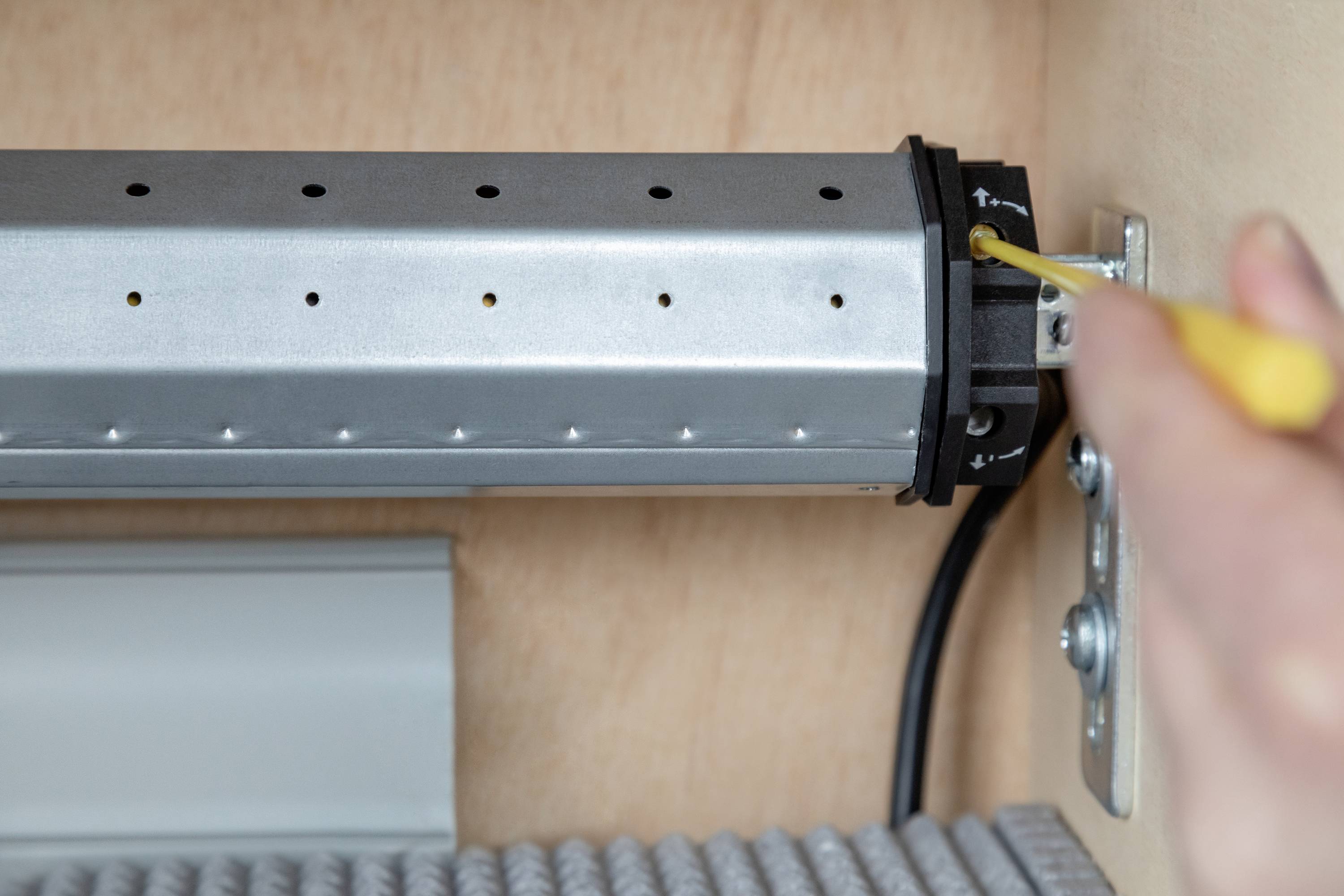 A person is tightening a screw on a metal mechanism with a yellow screwdriver. The mechanism is attached to a wooden board.