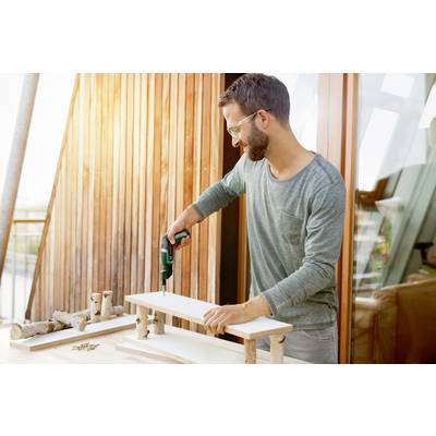 A man wearing safety glasses and a green jumper is assembling wooden pieces on a terrace using an electric screwdriver.