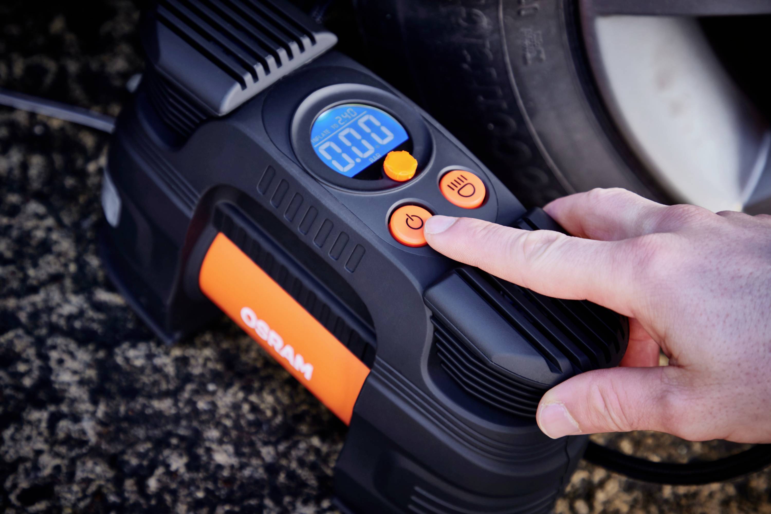 A person presses the power button of a digital tyre inflator lying next to a car tyre. The display shows '0.0'.