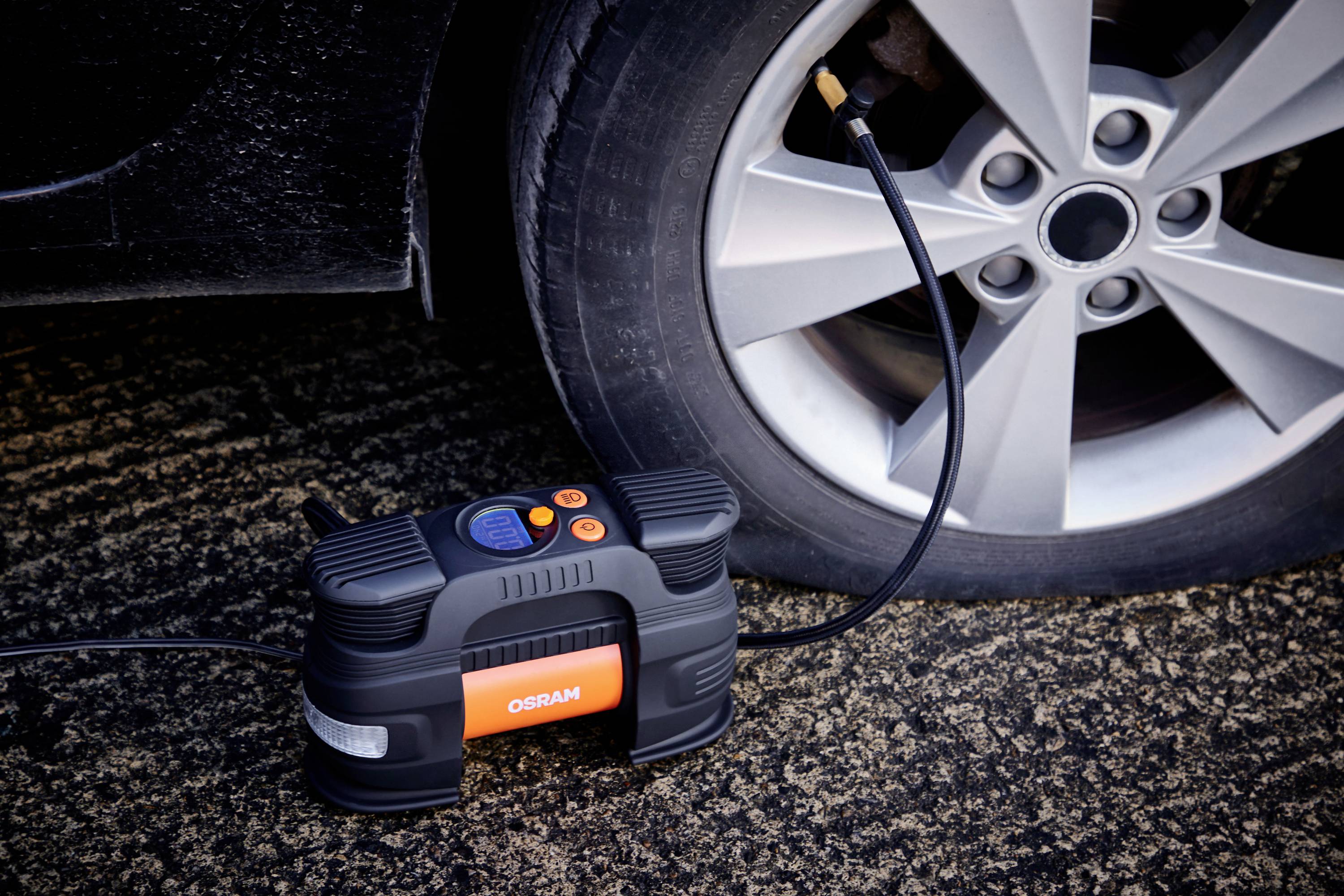 A portable air compressor is inflating a car tyre. The black compressor displays a digital readout and is switched on.