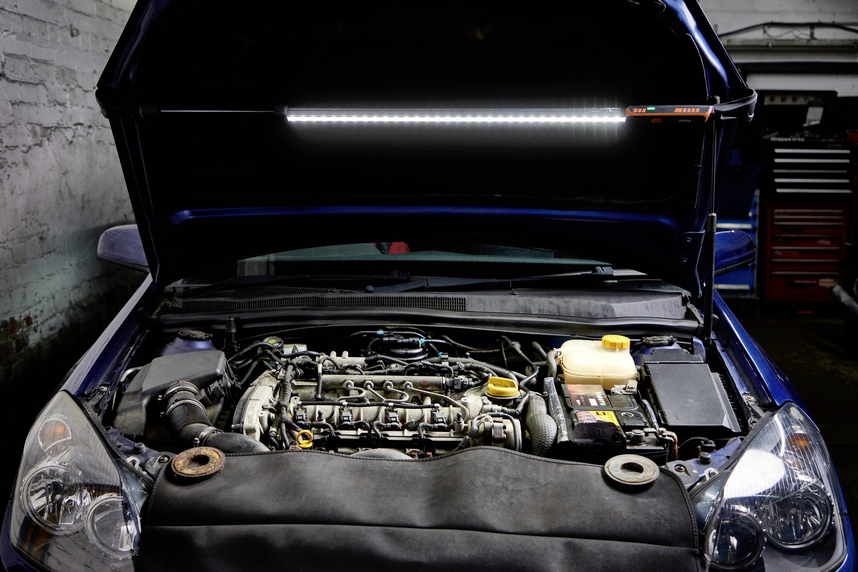Car with open bonnet in a workshop; engine block and tools visible, work light illuminating the engine.