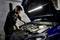 A mechanic repairs an engine beneath an open bonnet in a workshop, illuminated by an LED lamp.