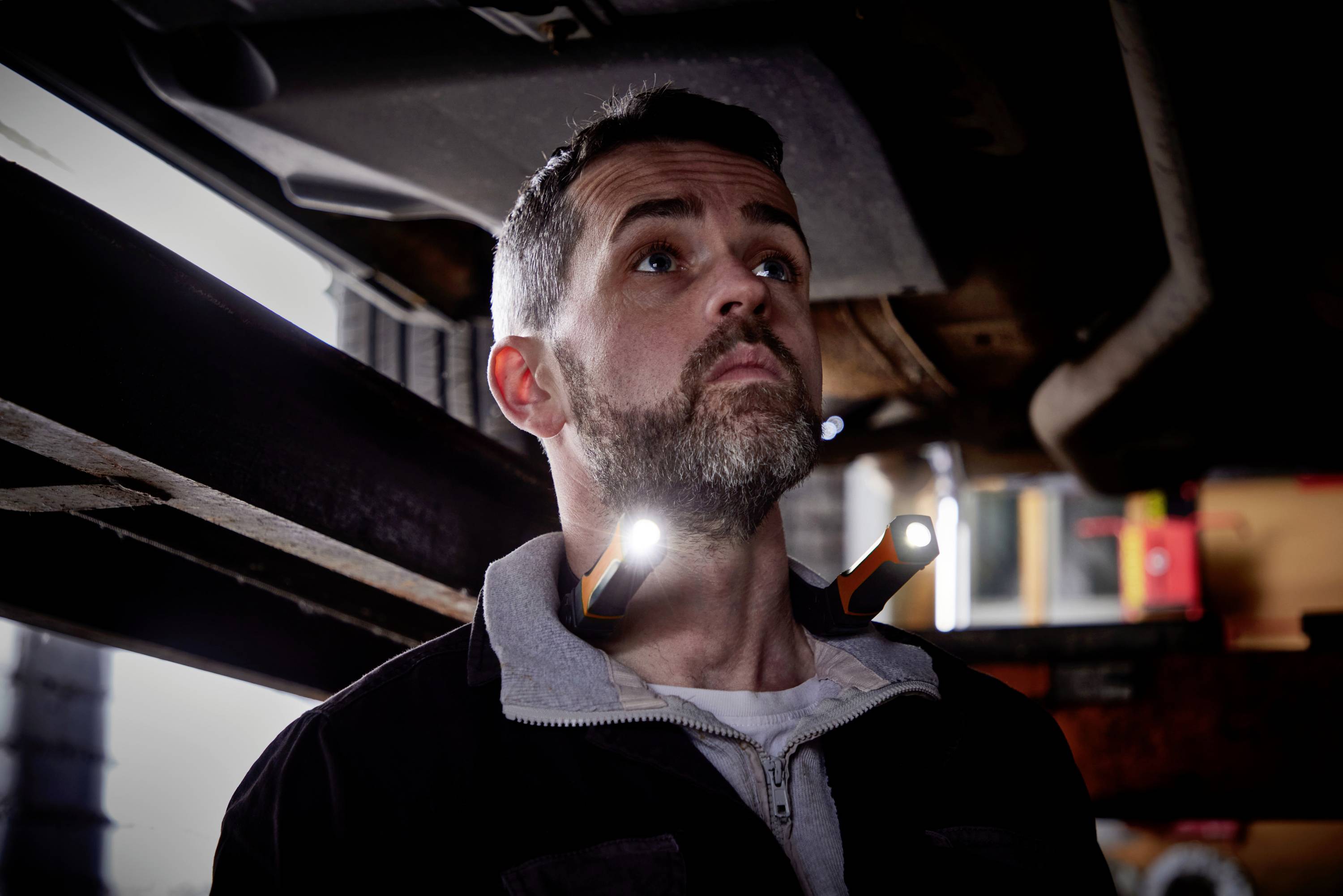 A man is working underneath a vehicle, illuminated by two small lights on his collar. He looks up with concentration.