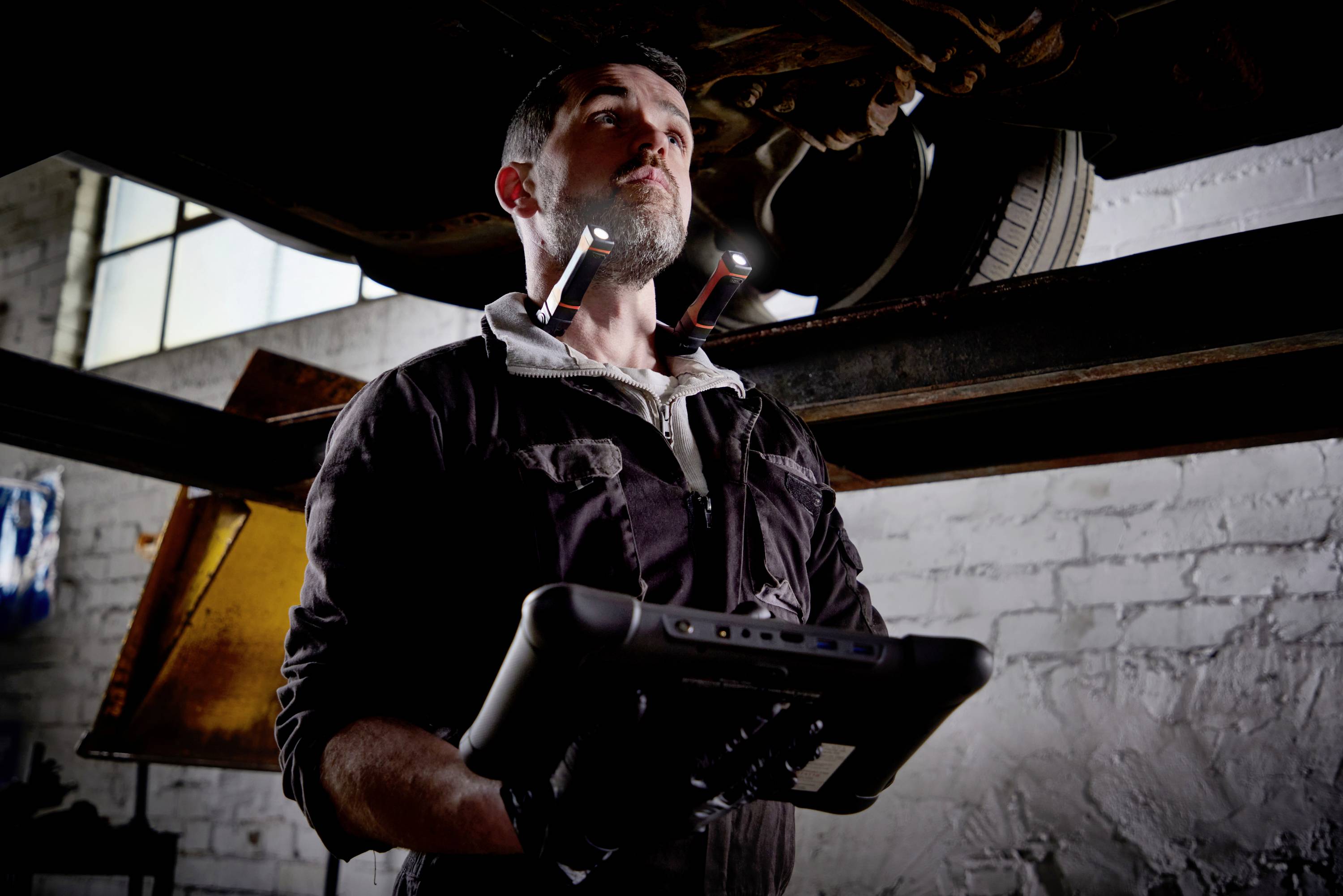 A mechanic inspects a vehicle in a workshop, holding a diagnostic device in his hands while looking underneath the car.