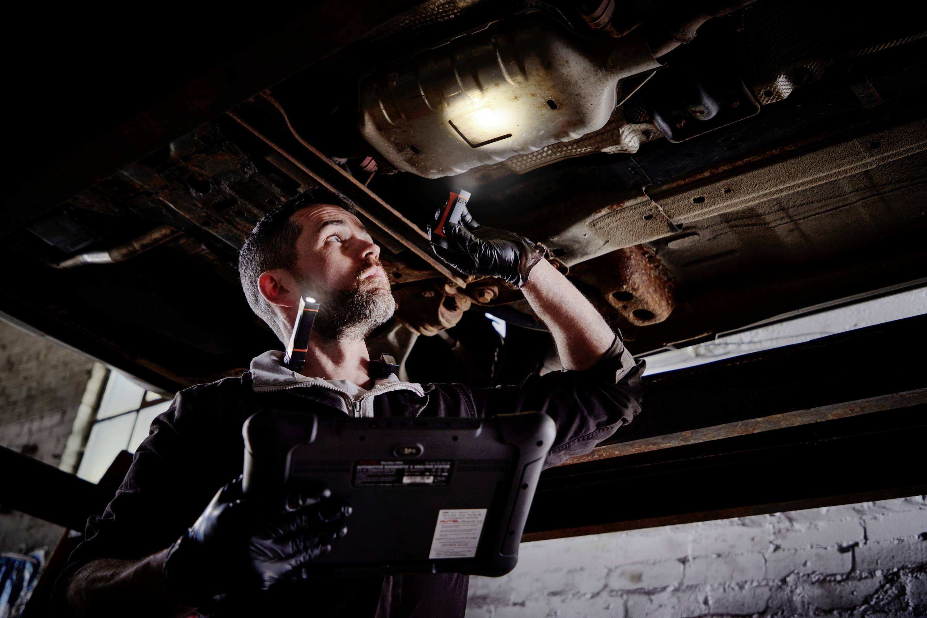A mechanic is inspecting the underside of a car in a garage. He is using a torch and holding a diagnostic device.