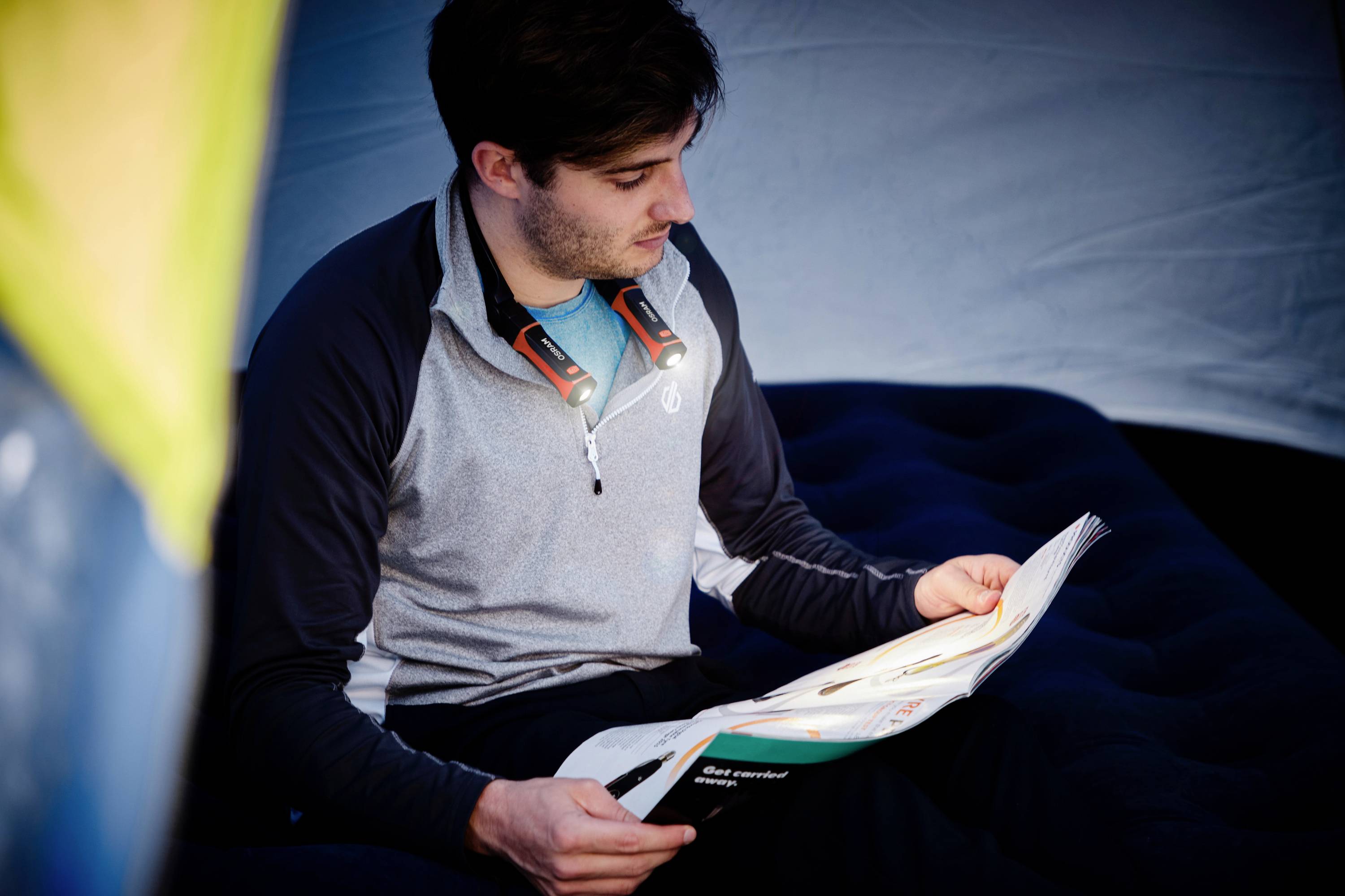 A person is sitting in a tent and reading a magazine. They are wearing a lamp around their neck to make reading easier.