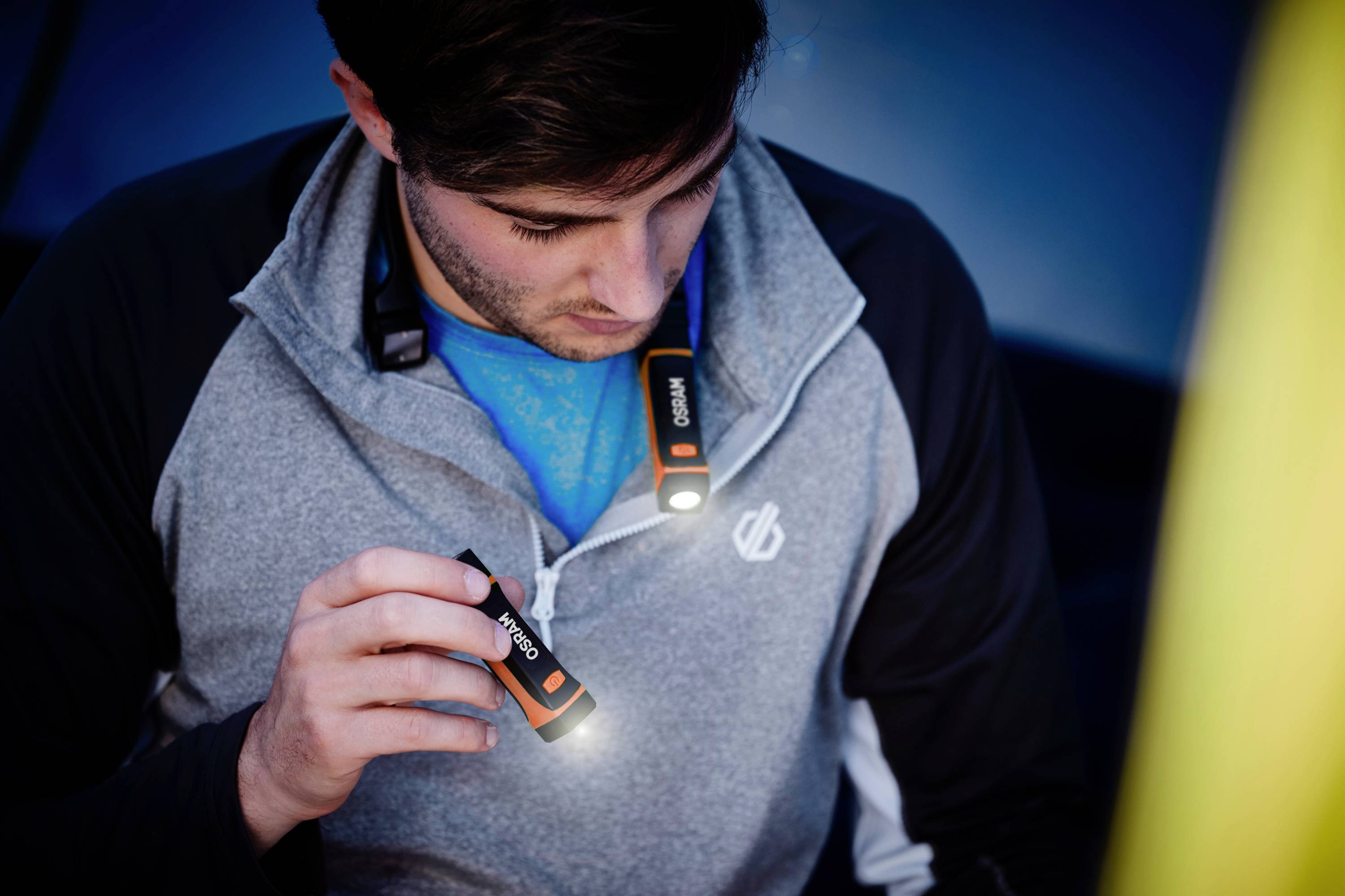 A man is holding a small torch that is illuminating the space while he sits inside a tent. He is wearing a grey jumper.