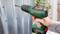 A person is drilling a hole in a corrugated sheet metal using a green Bosch cordless drill. A garden is blurrily visible in the background.