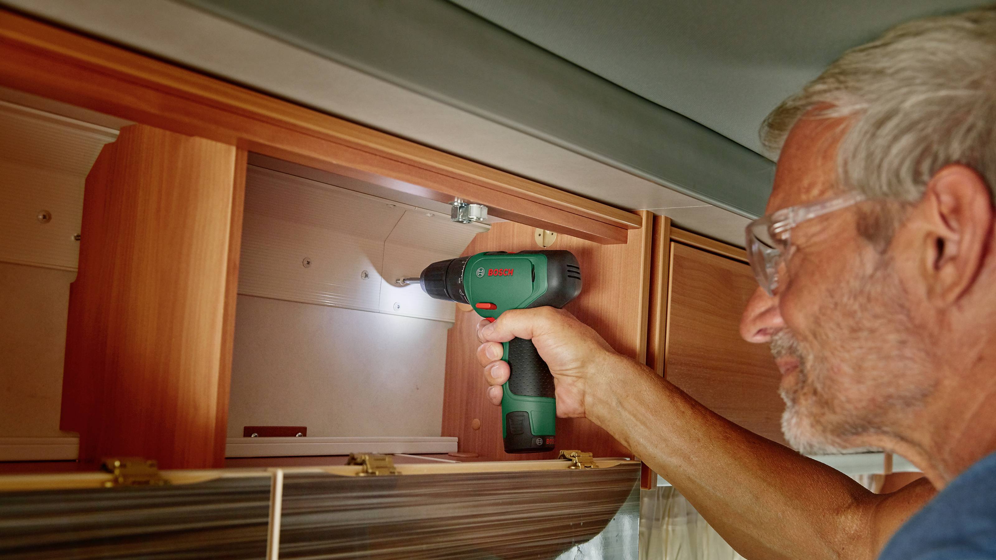 A man is using a cordless drill driver to secure a shelf in a motorhome. He is wearing safety glasses.