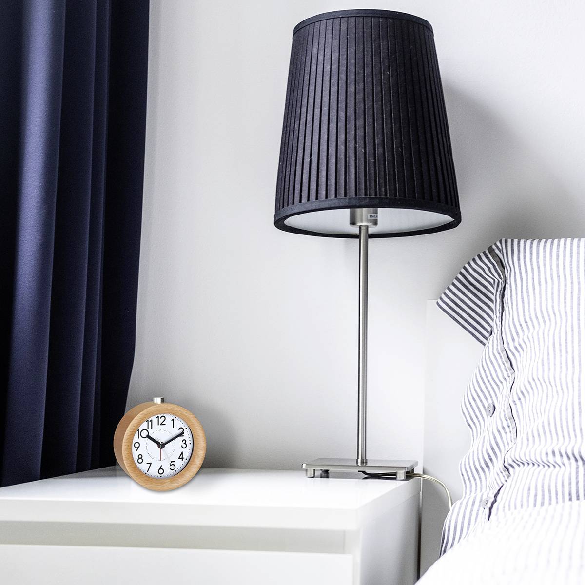 On a bedside table, there is a black table lamp and an alarm clock with a brown frame. Next to it is a bed with a striped duvet.