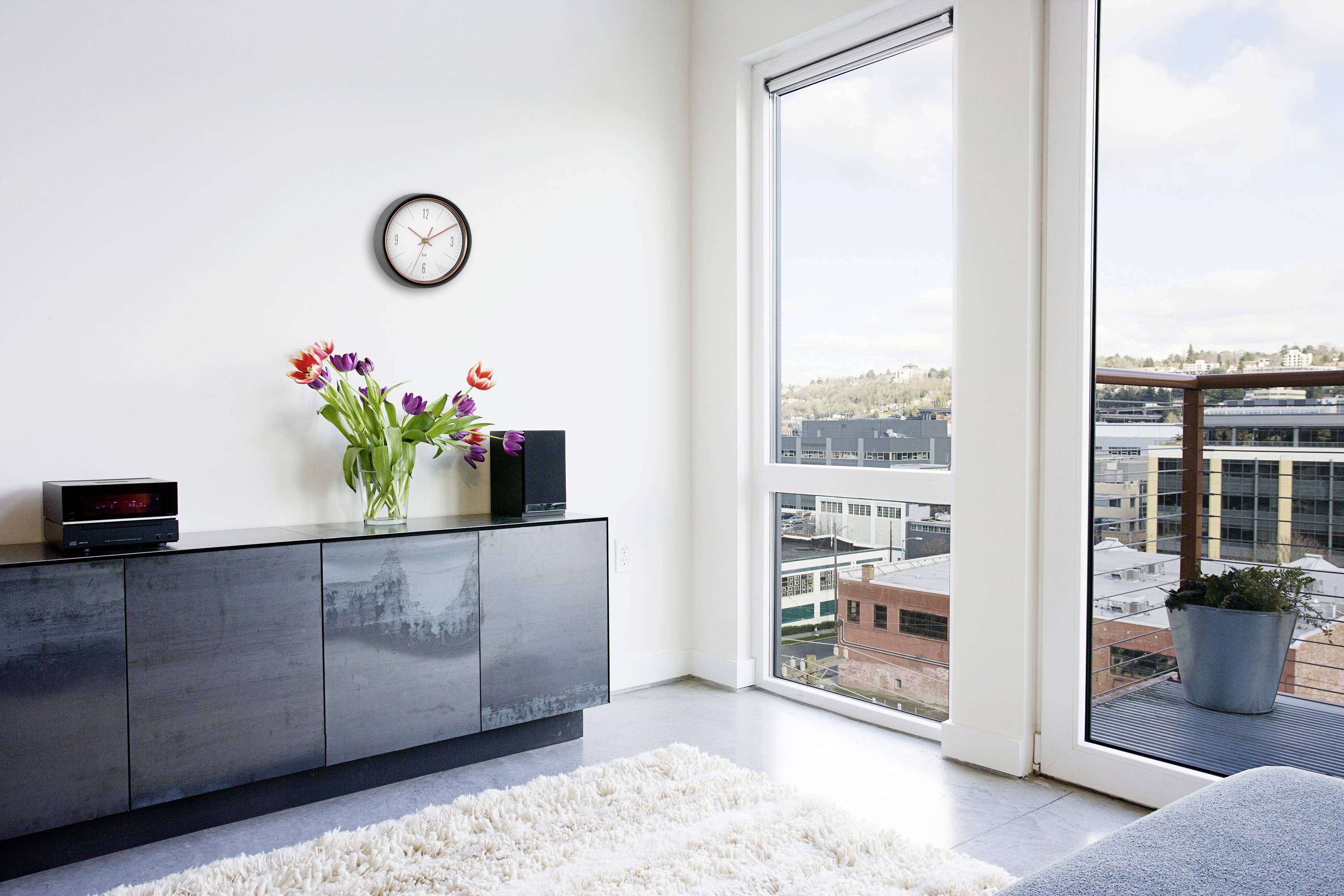 Modern living room view with a clear window view of city buildings, wall clock, sideboard with a vase of colourful flowers.