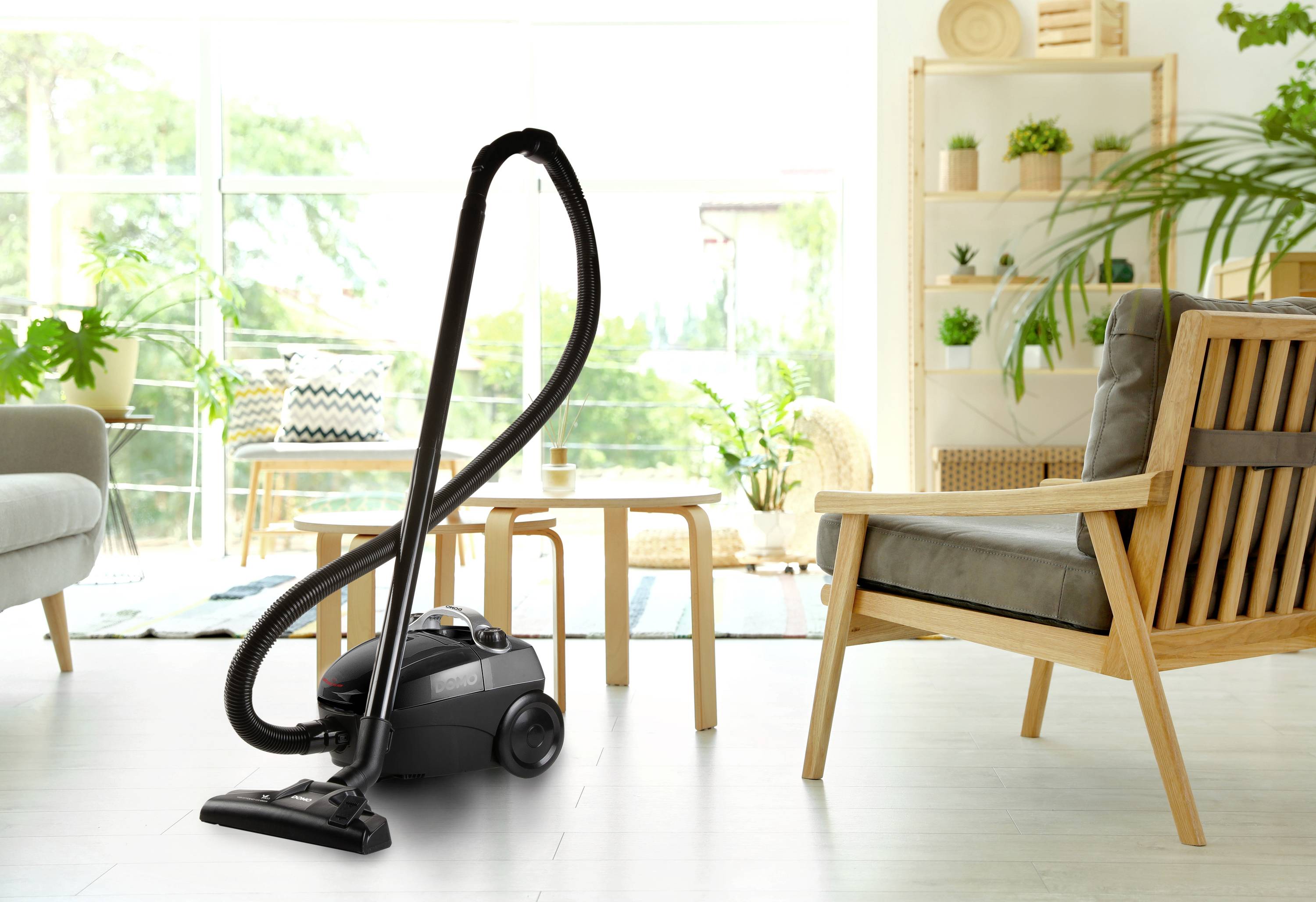 Vacuum cleaner stands in a modern living room with wooden furniture and plants, on a light wooden floor, in front of large windows.