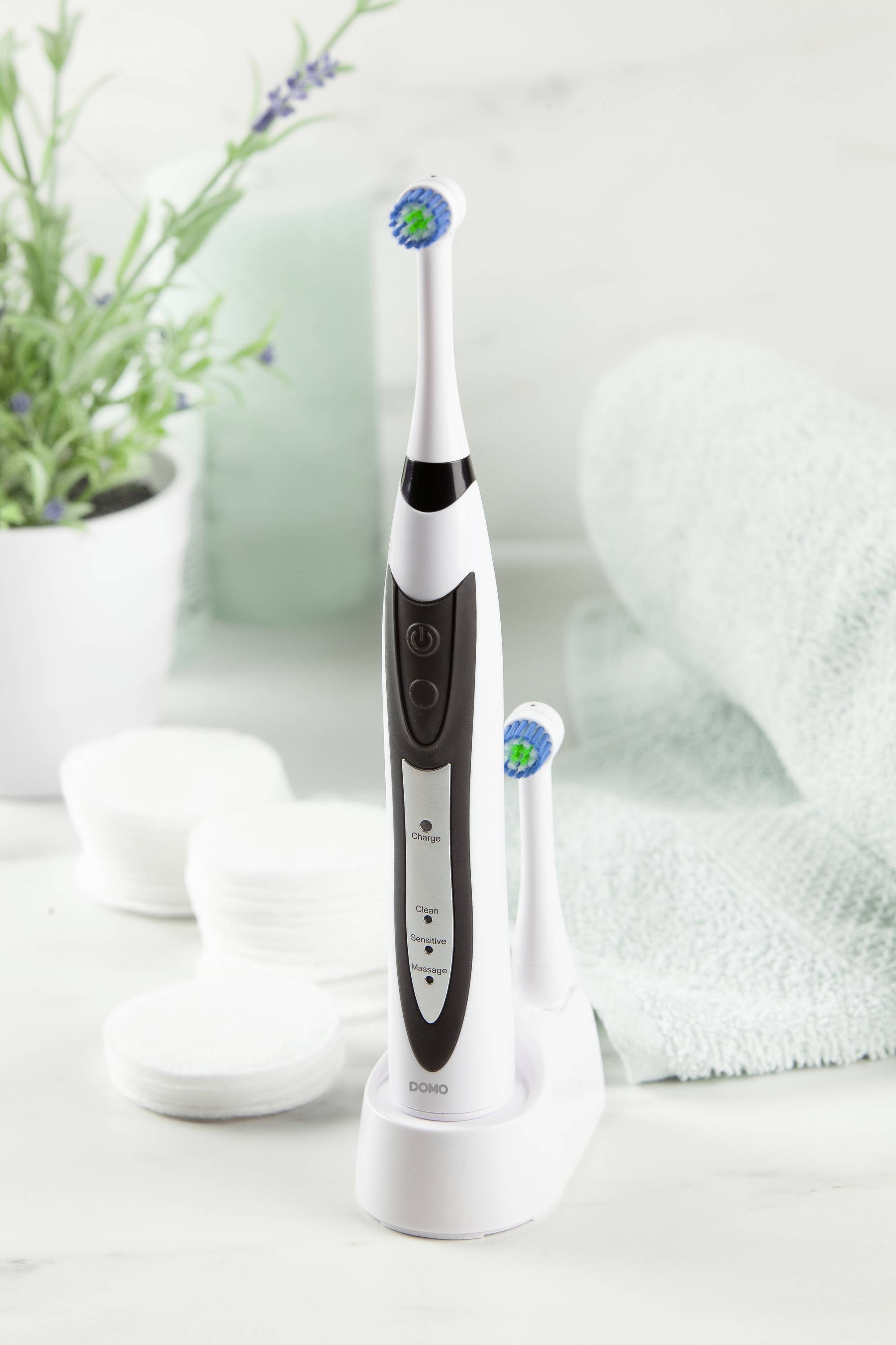 Electric toothbrush on charging station, next to cotton pads and a towel, with a plant in the background.