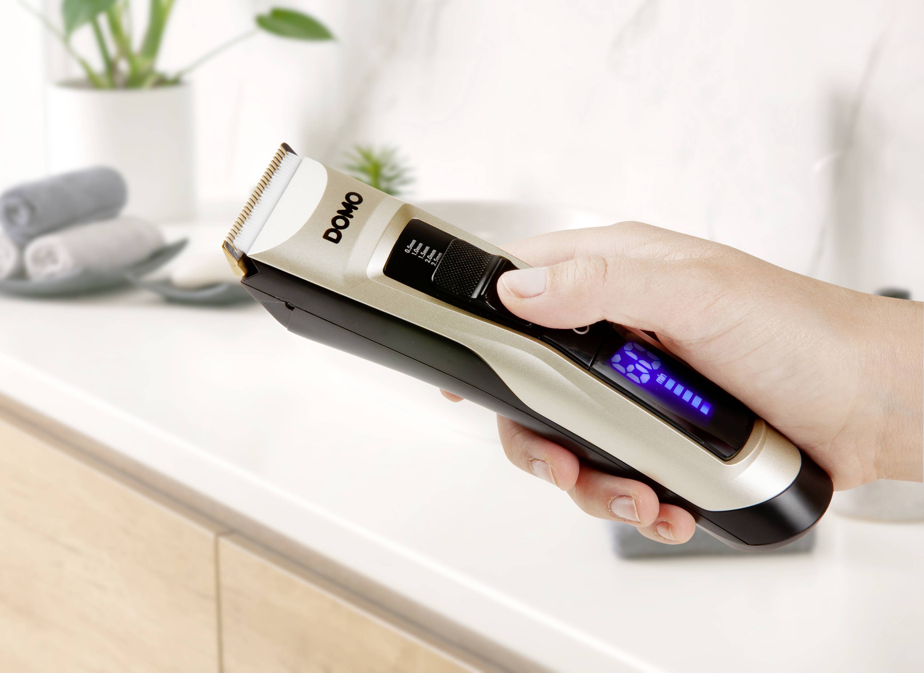 A hand is holding an electric hair clipper over a sink. The razor shows a blue LED display.