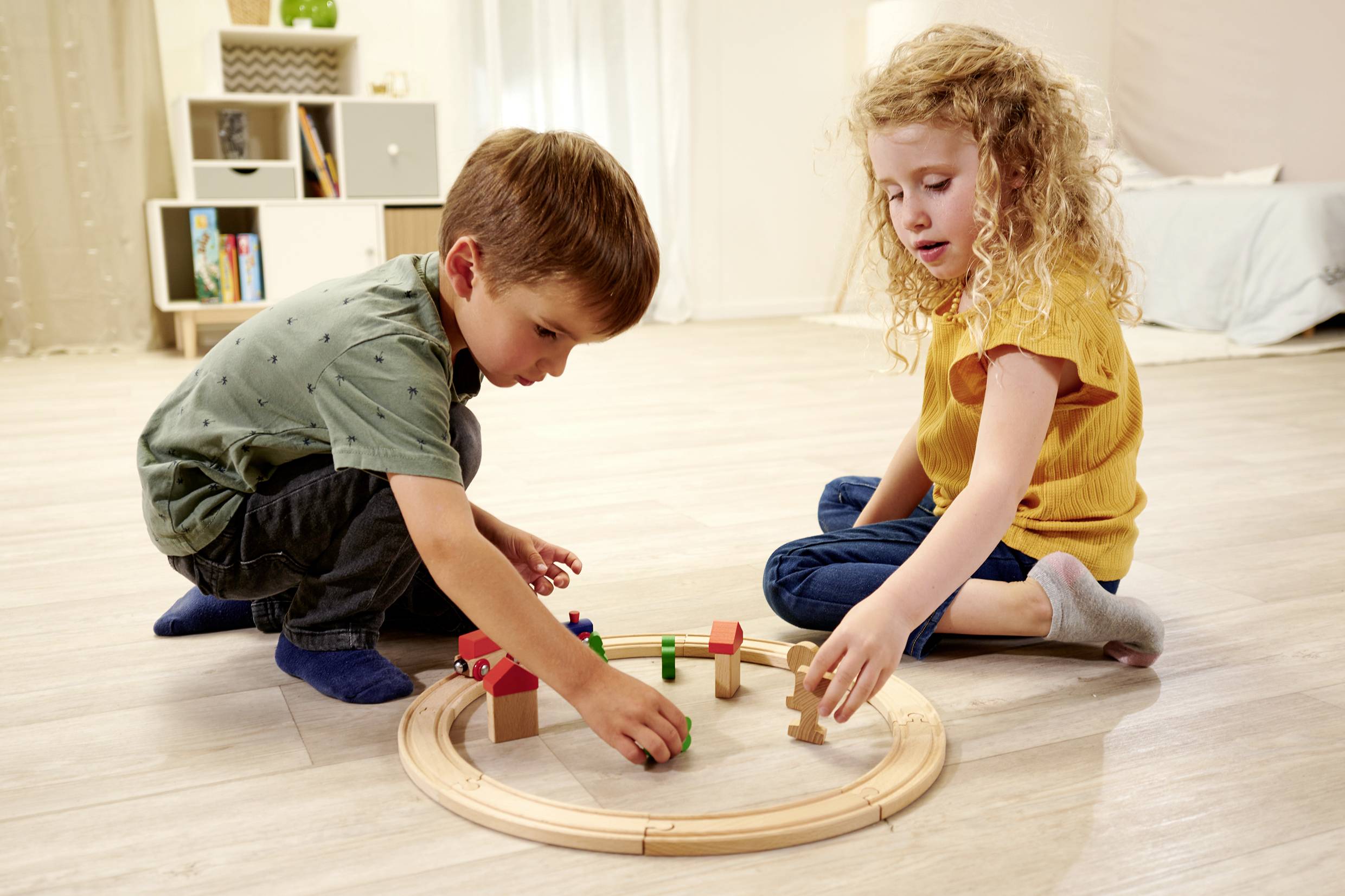 Two children are sitting on the floor and playing with a wooden toy train and building blocks in a brightly lit room.