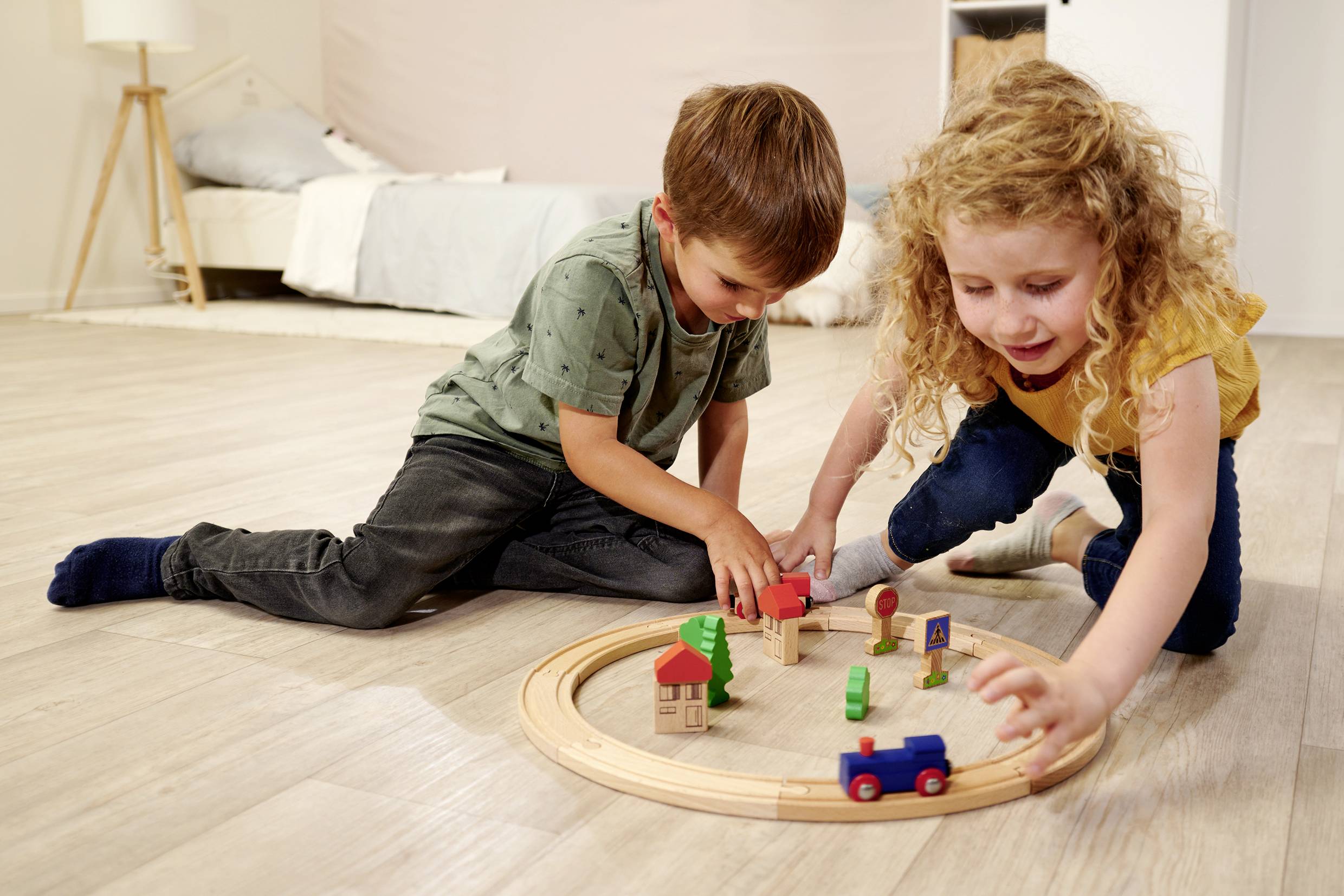 Two children are playing with a wooden train set on the floor of a children's bedroom. In the background, a bed is visible.