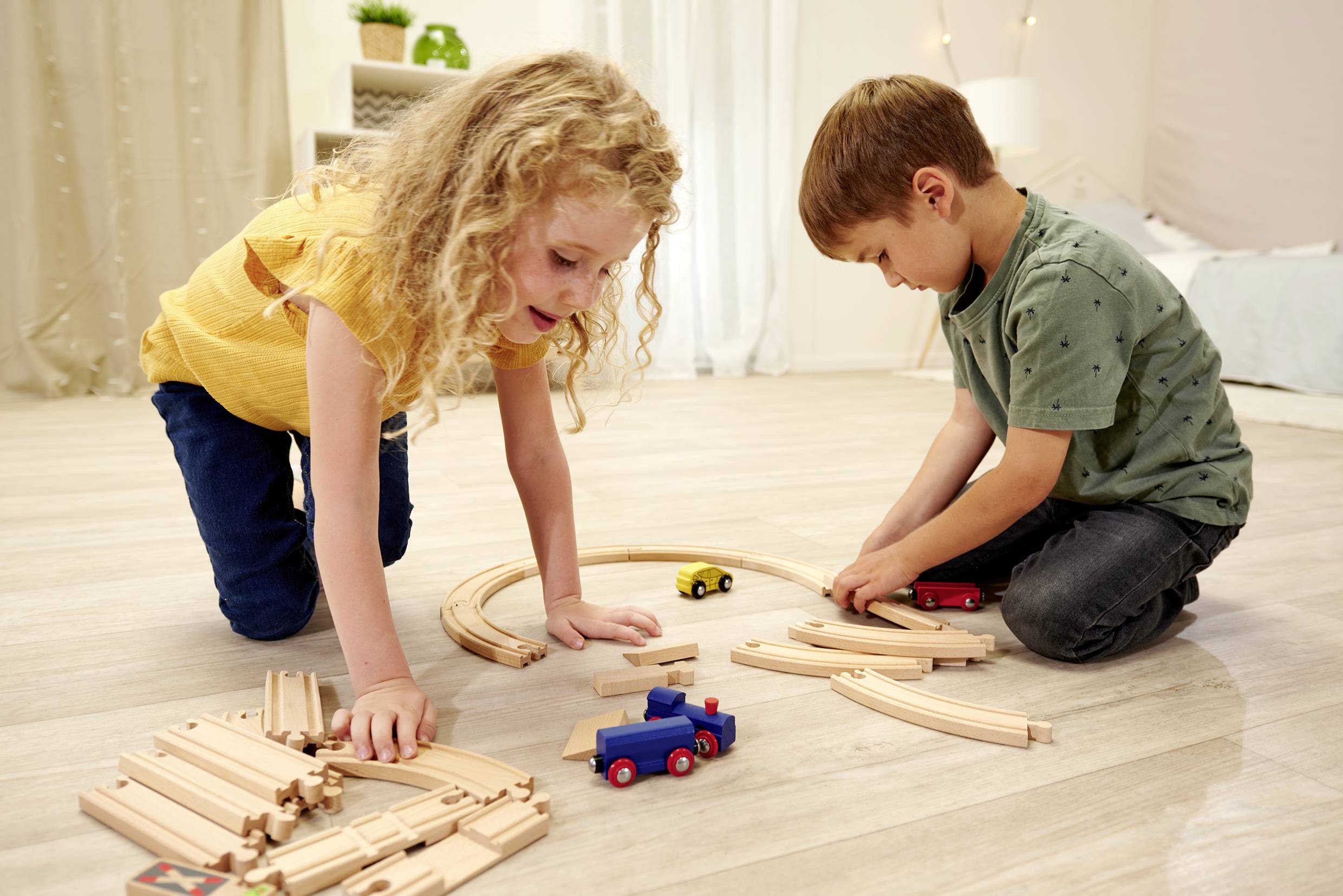 Two children are playing on the floor with a wooden train set. A girl and a boy are concentrating on assembling the tracks.