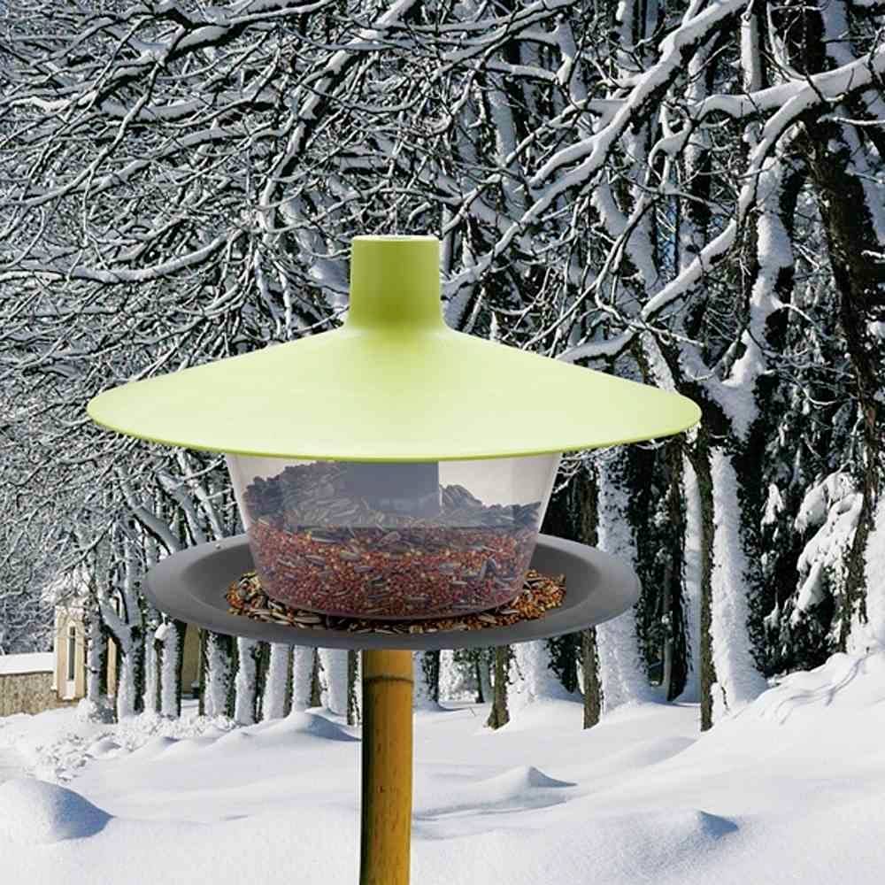 A bird feeder with a green cover, filled with seeds, stands in a snowy winter forest with snow-covered trees in the background.