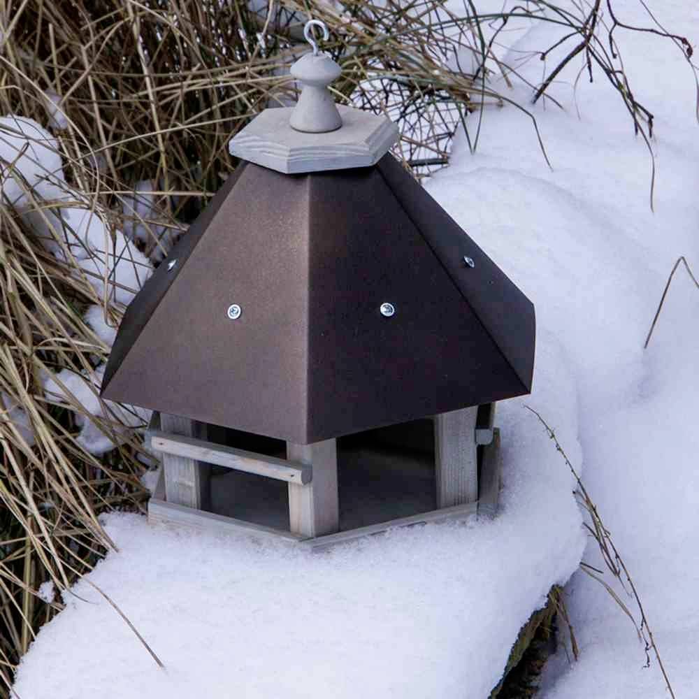 Wooden bird feeder with a hexagonal roof, standing on snow. Surrounded by tall, dry grasses protruding from the snow.
