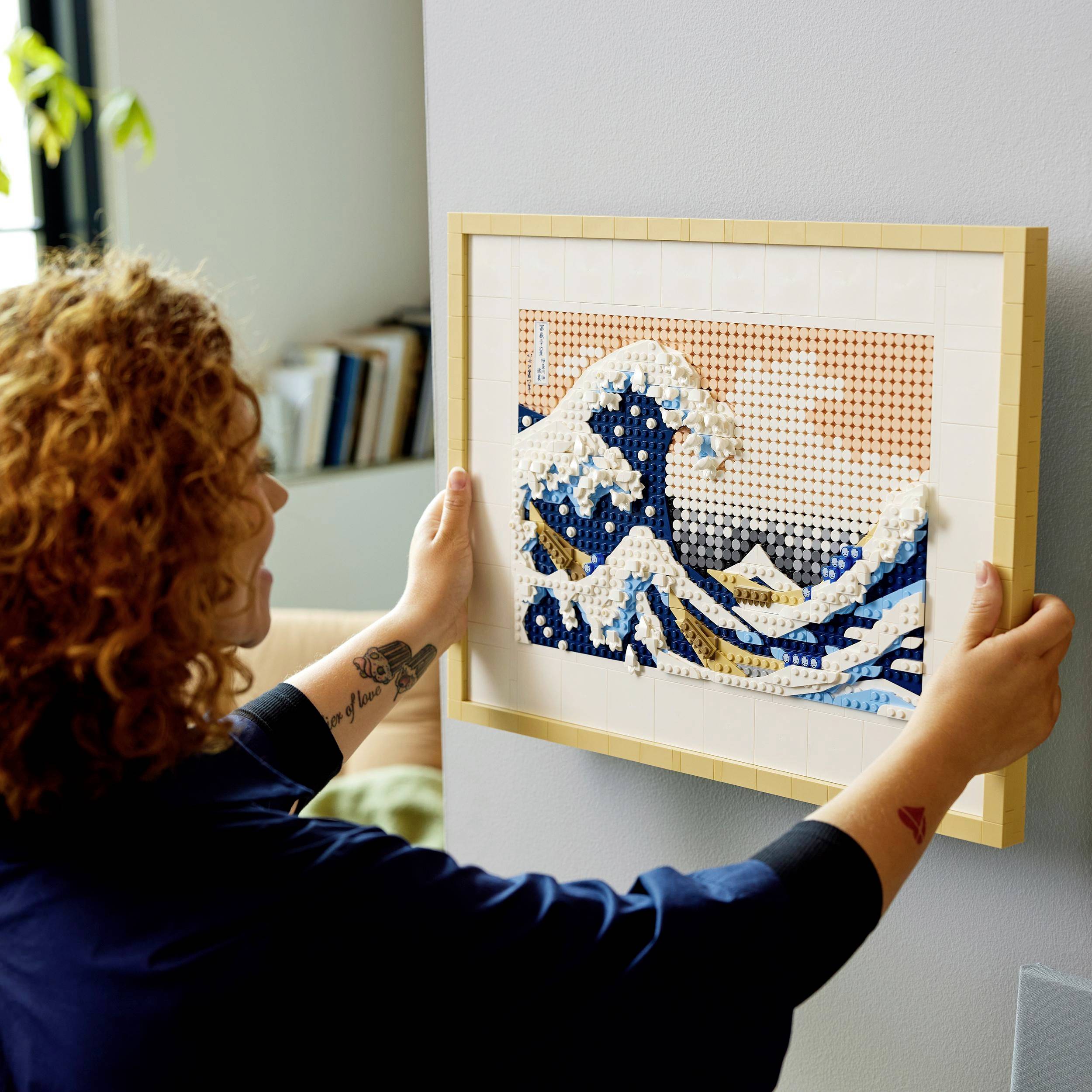 A person is hanging a framed mosaic of the 'Great Wave off Kanagawa' on a wall. The mosaic is composed of colourful, pixel-like elements.
