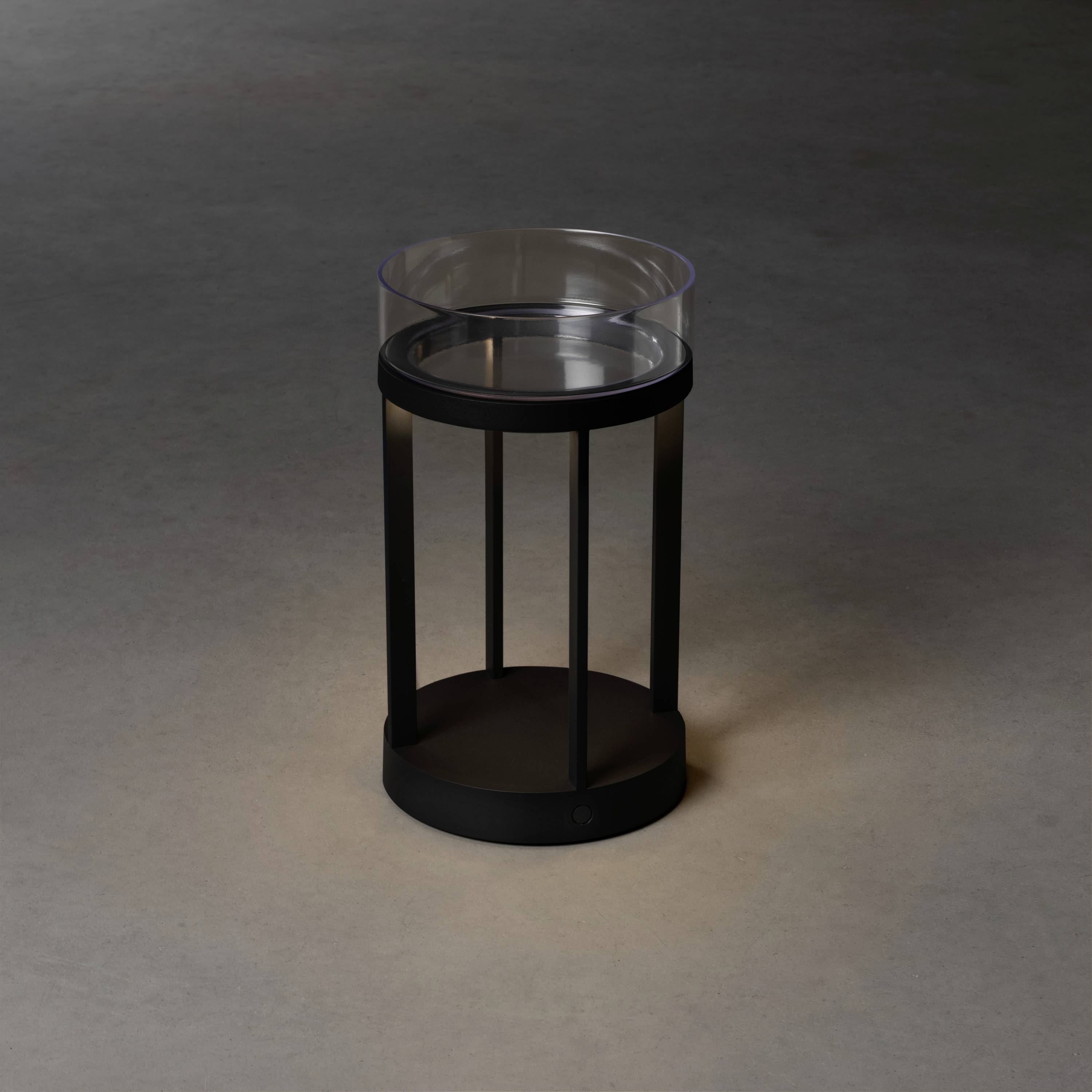 A round, contemporary side table made of black metal with a transparent glass top sits on a grey floor.
