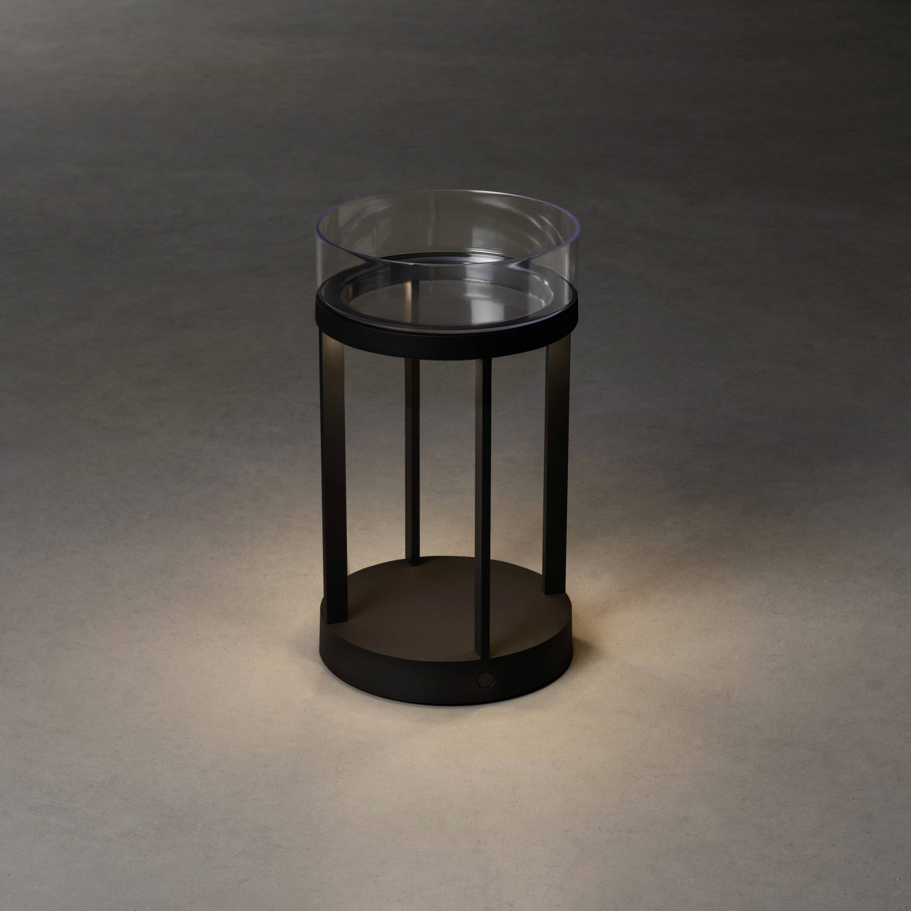 A round table with a transparent glass top and black metal legs, illuminated by soft light in a dark room.