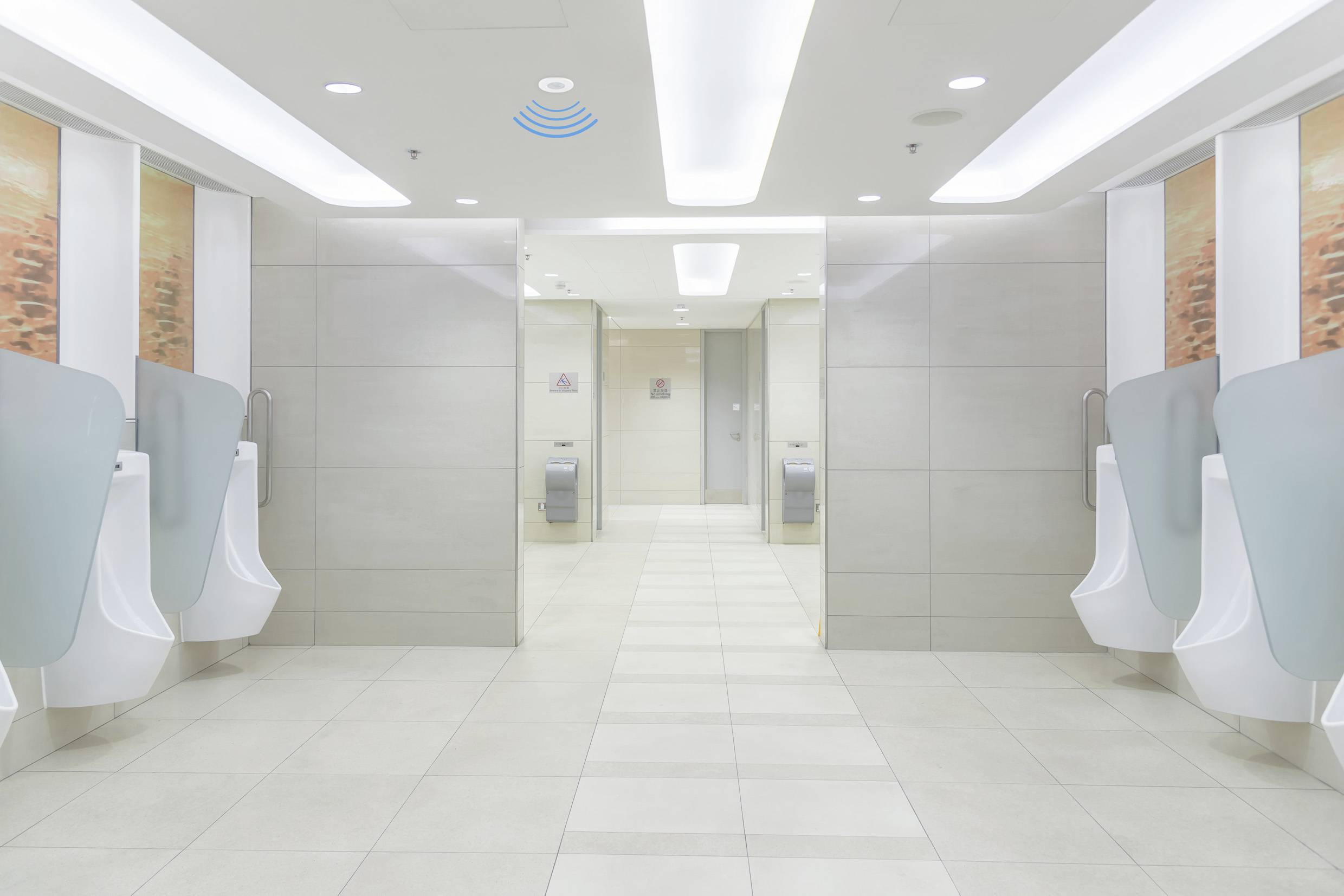 A vacant, bright corridor in a public lavatory with urinals along both walls and bins located at the rear of the space.