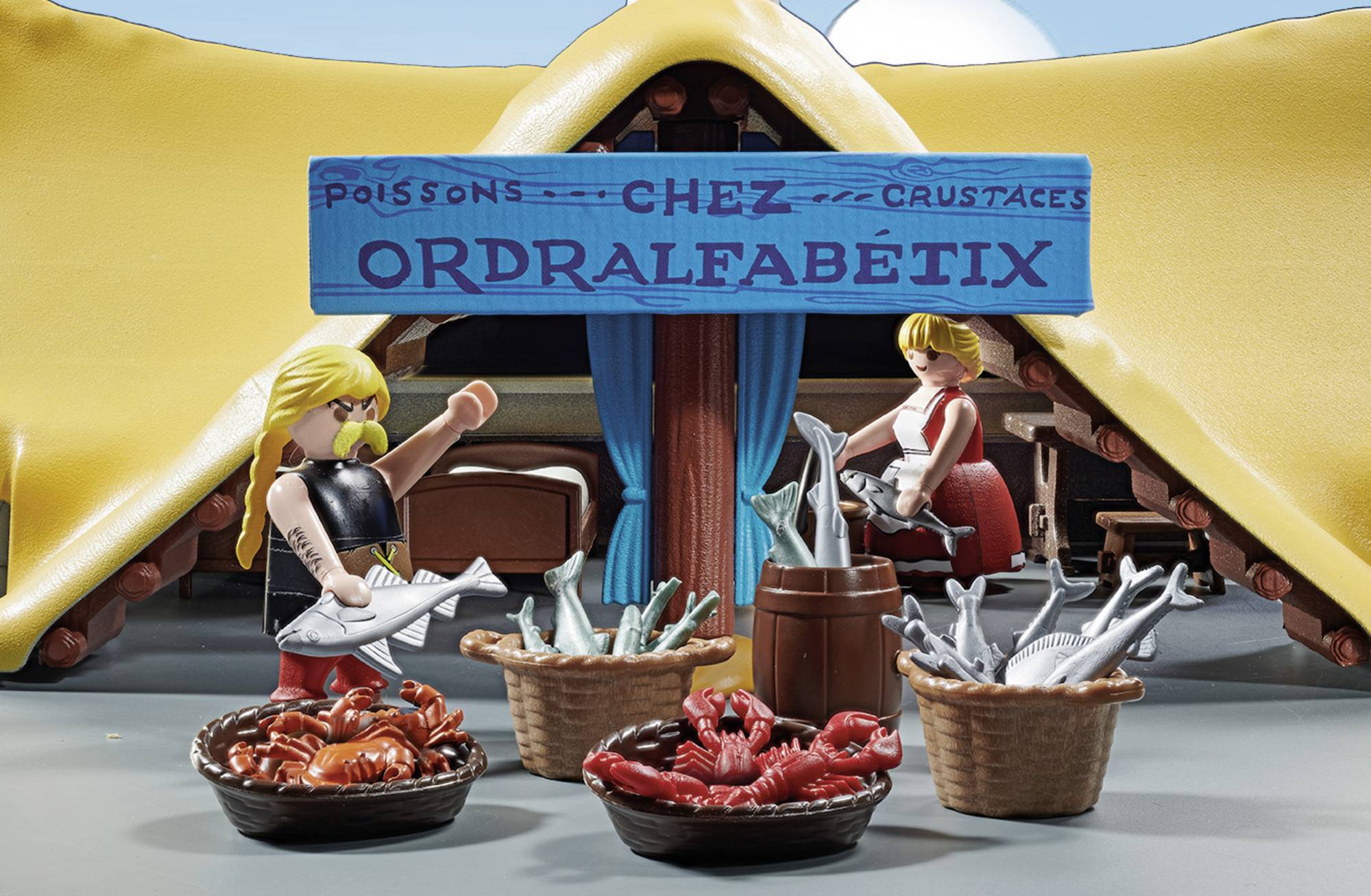 Two figures in front of a fish shop 'Chez Ordralfabetix' with baskets full of fish and crustaceans. Figure on the left is holding up a fish.