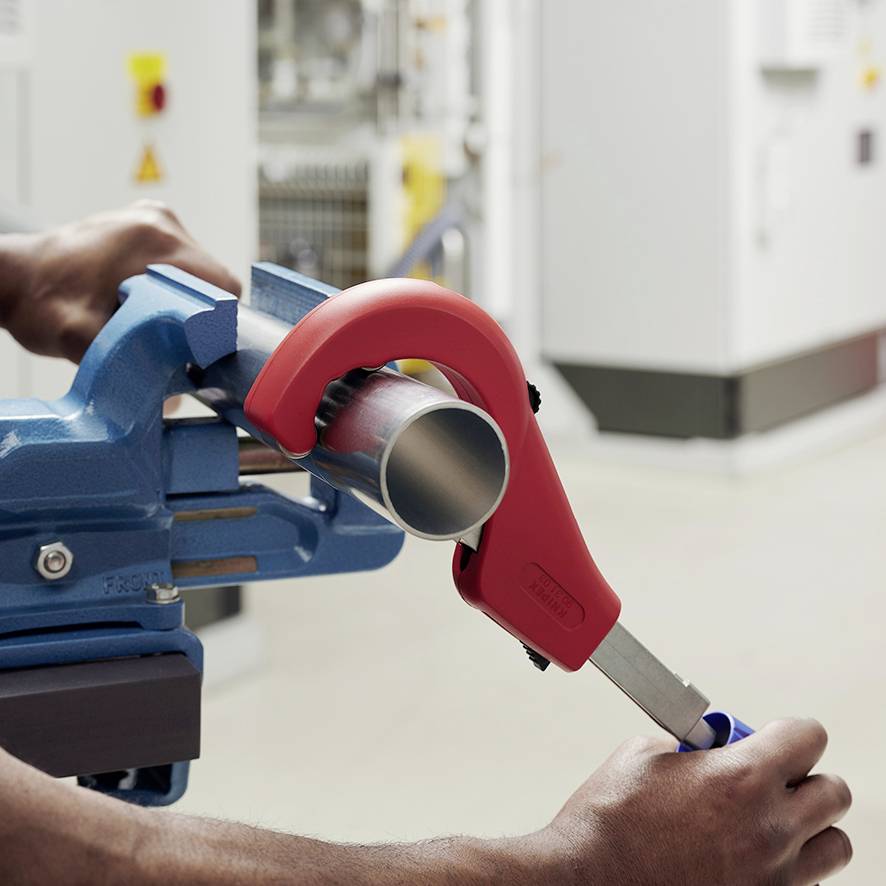 A person is cutting a metal pipe with a red pipe cutter, the pipe is clamped in a blue vice.