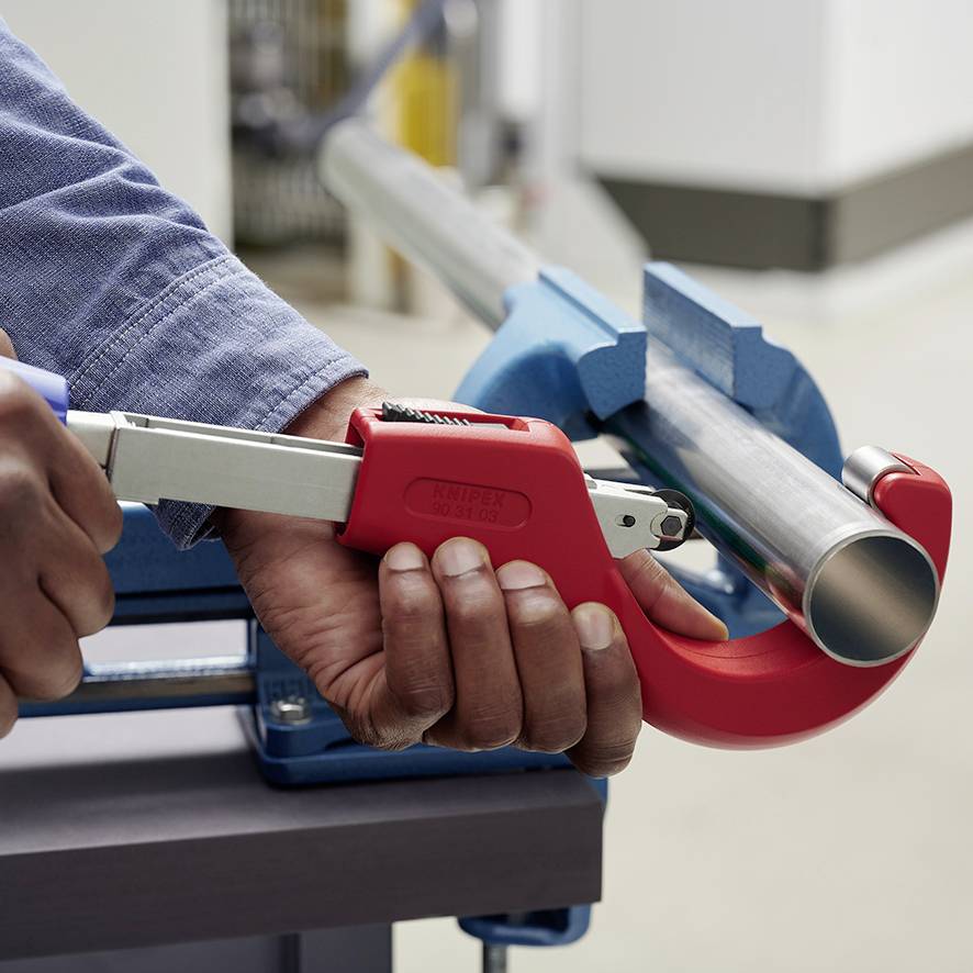 A person is cutting a metal pipe with a red pipe cutter. The focus is on the hand and the cutting tool.