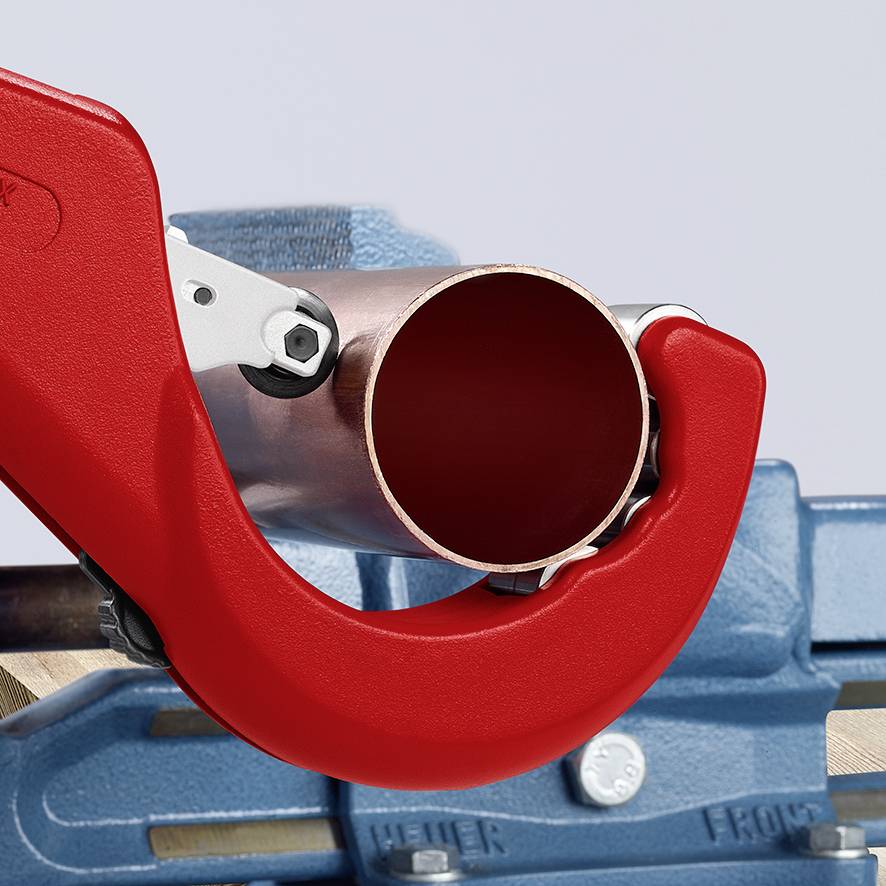 Rohrschneider cuts a copper pipe; metal pipe is clamped in a red blade. Background shows a workbench and tools.