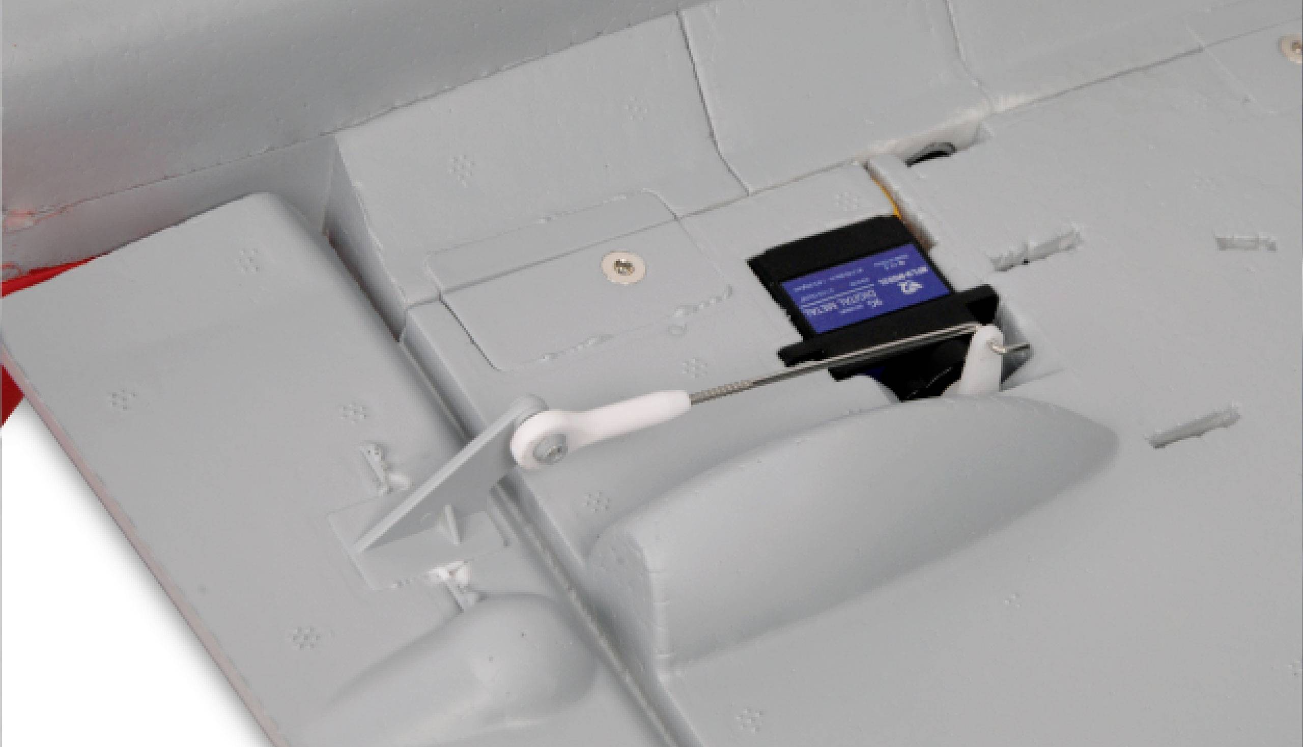 Close-up of a white model aircraft with a blue servo in the wing. The servo controls a control surface movement via a metal rod.