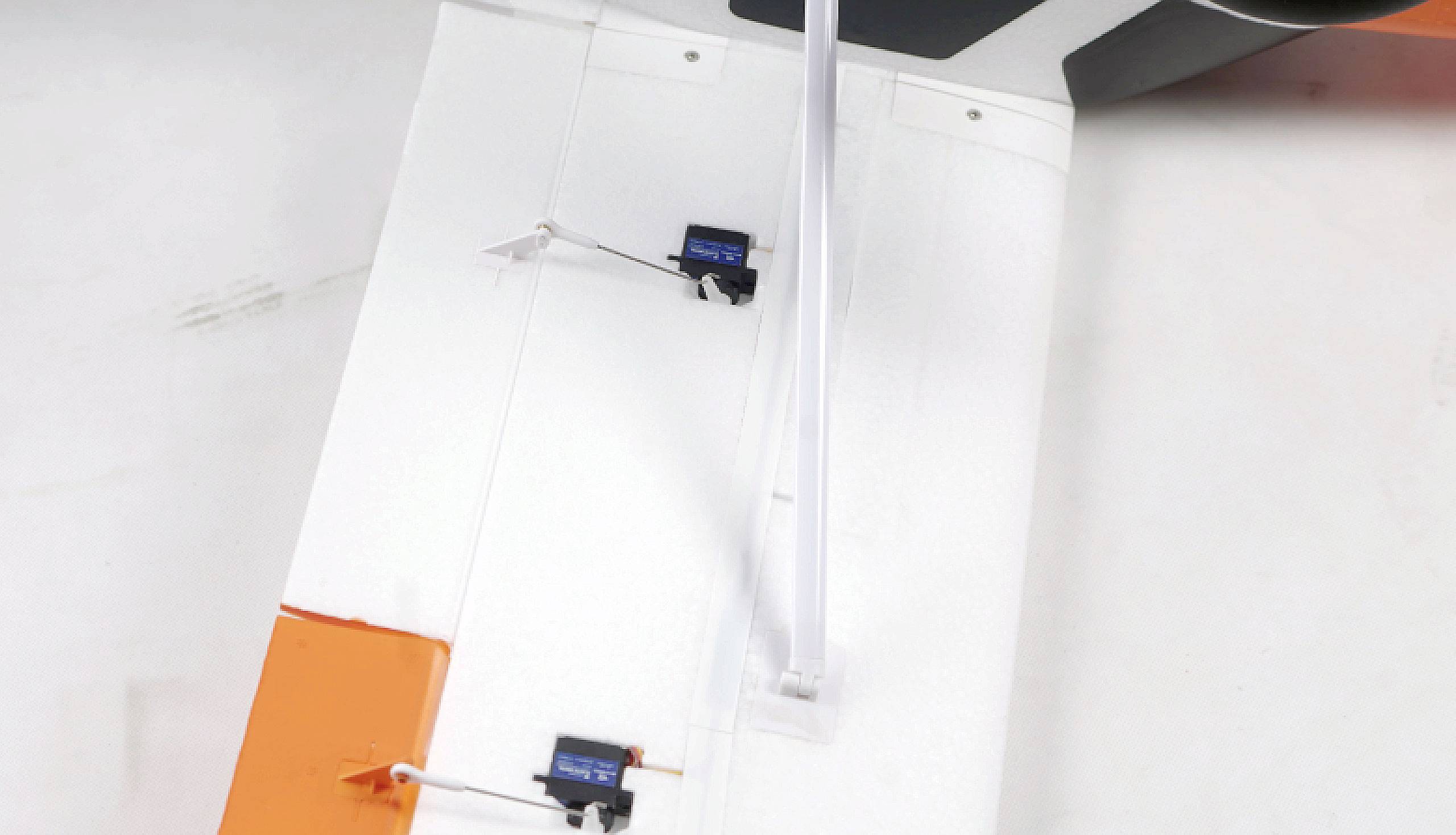 A model aircraft wing with orange and white paintwork, showing two integrated servos with control rods for wing control.