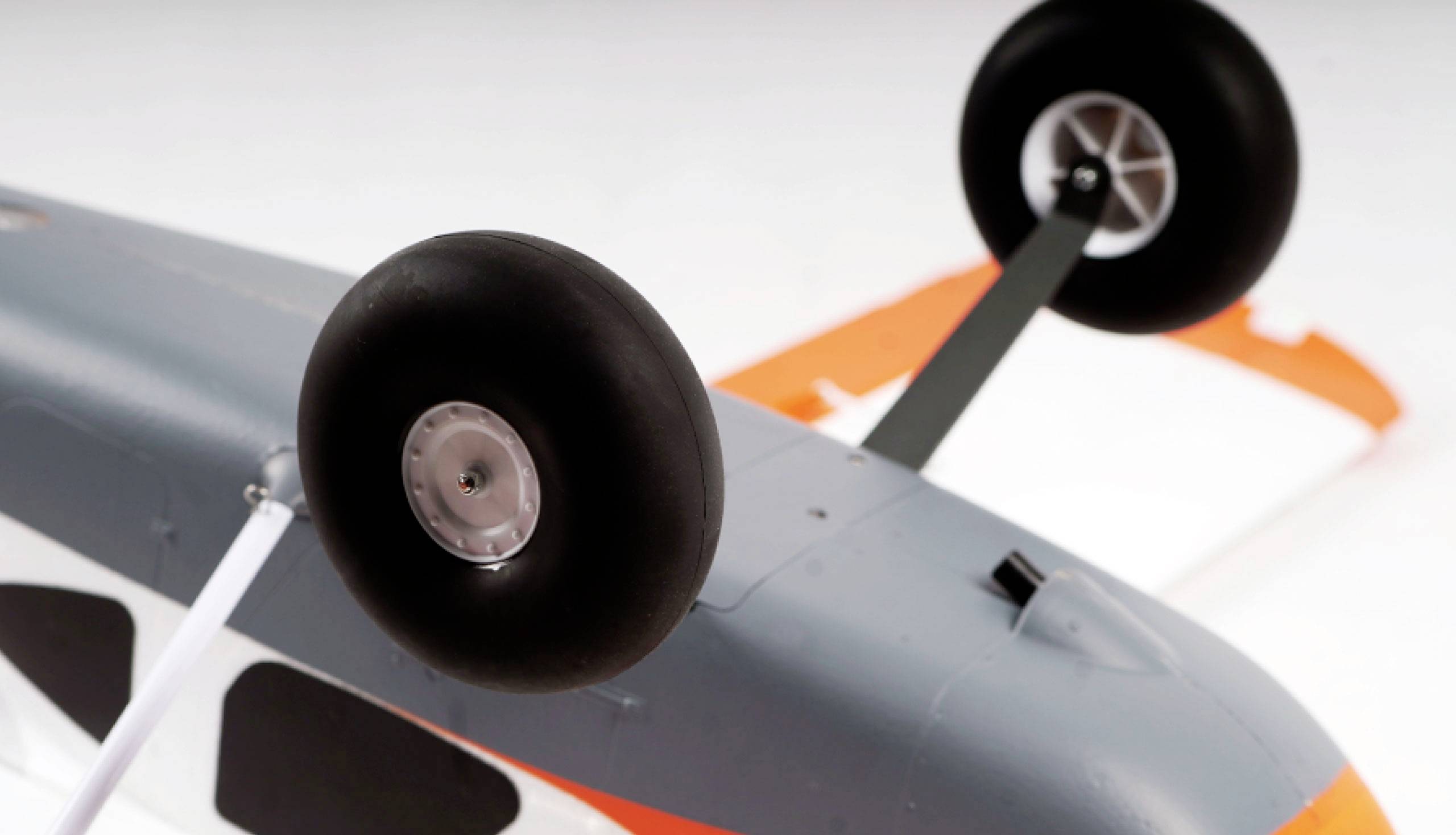 Close-up of a model aircraft fuselage positioned upside down. The landing gear wheels are visible, with a grey-white fuselage colour and part of an orange wing.