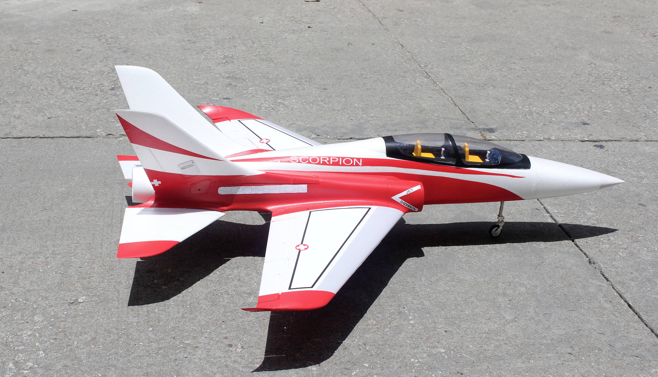 Model aircraft in red and white on an asphalt surface. It has a sleek design with a double-seat cockpit and visible wings and tail surfaces.