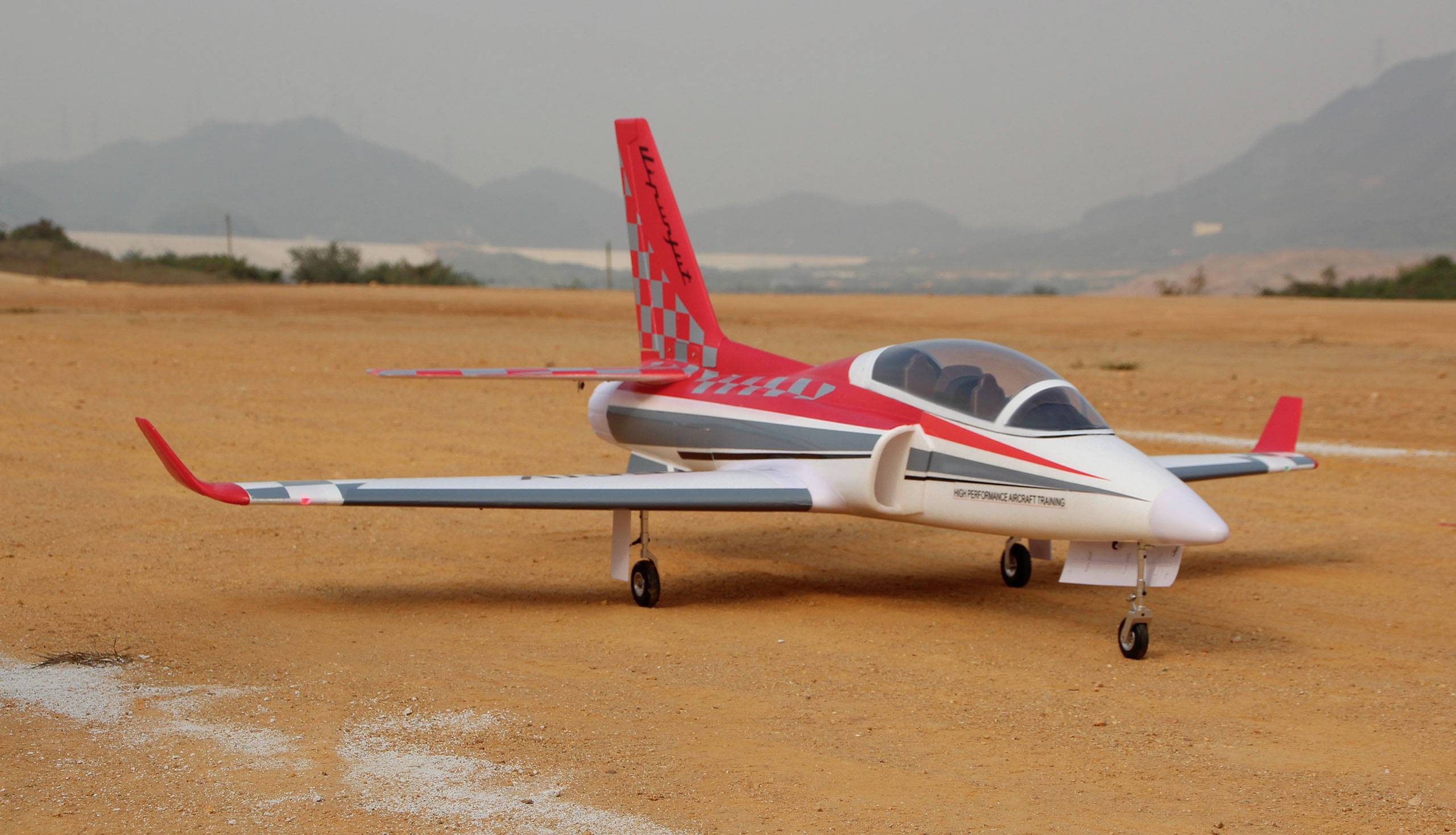 A remote-controlled model aircraft with red and white design is standing on a sandy airfield, surrounded by mountains in the background.