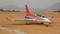 A remote-controlled model aircraft with red and white design is standing on a sandy airfield, surrounded by mountains in the background.