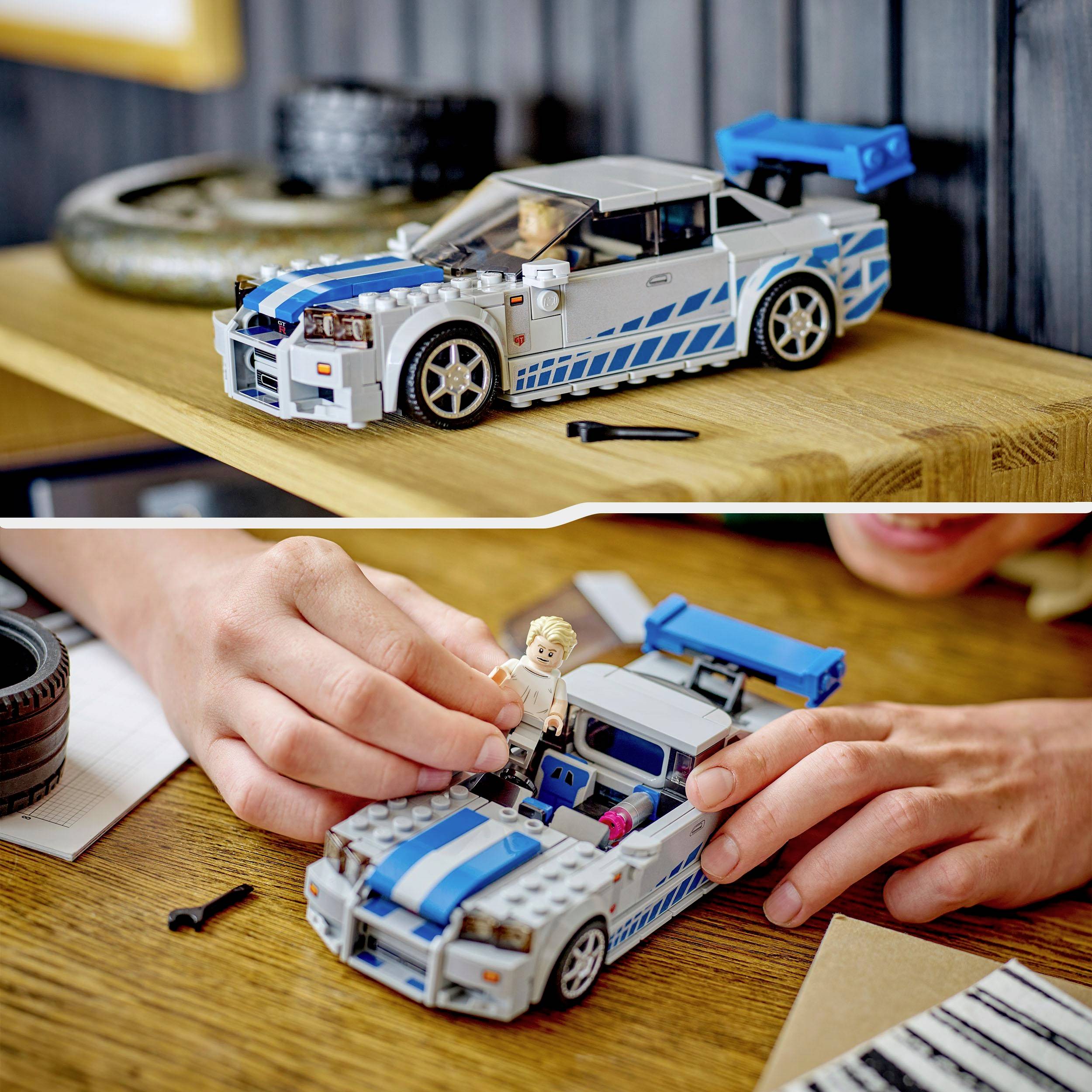 A model car made of building blocks with blue and grey paintwork is being assembled. A hand is placing a figurine into the car.